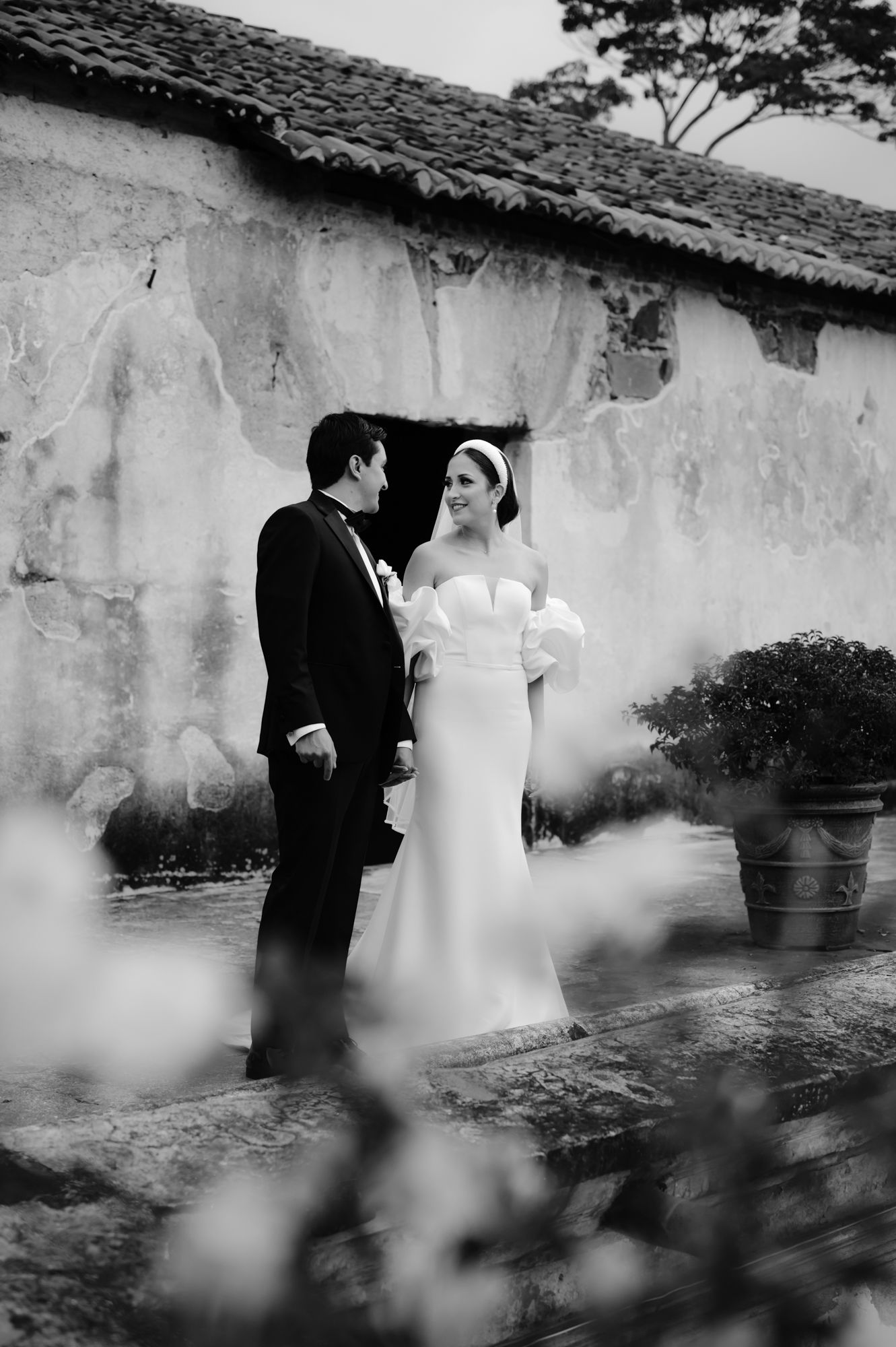 Black and white portrait of a newlywed couple standing against a weathered wall in an outdoor garden setting.