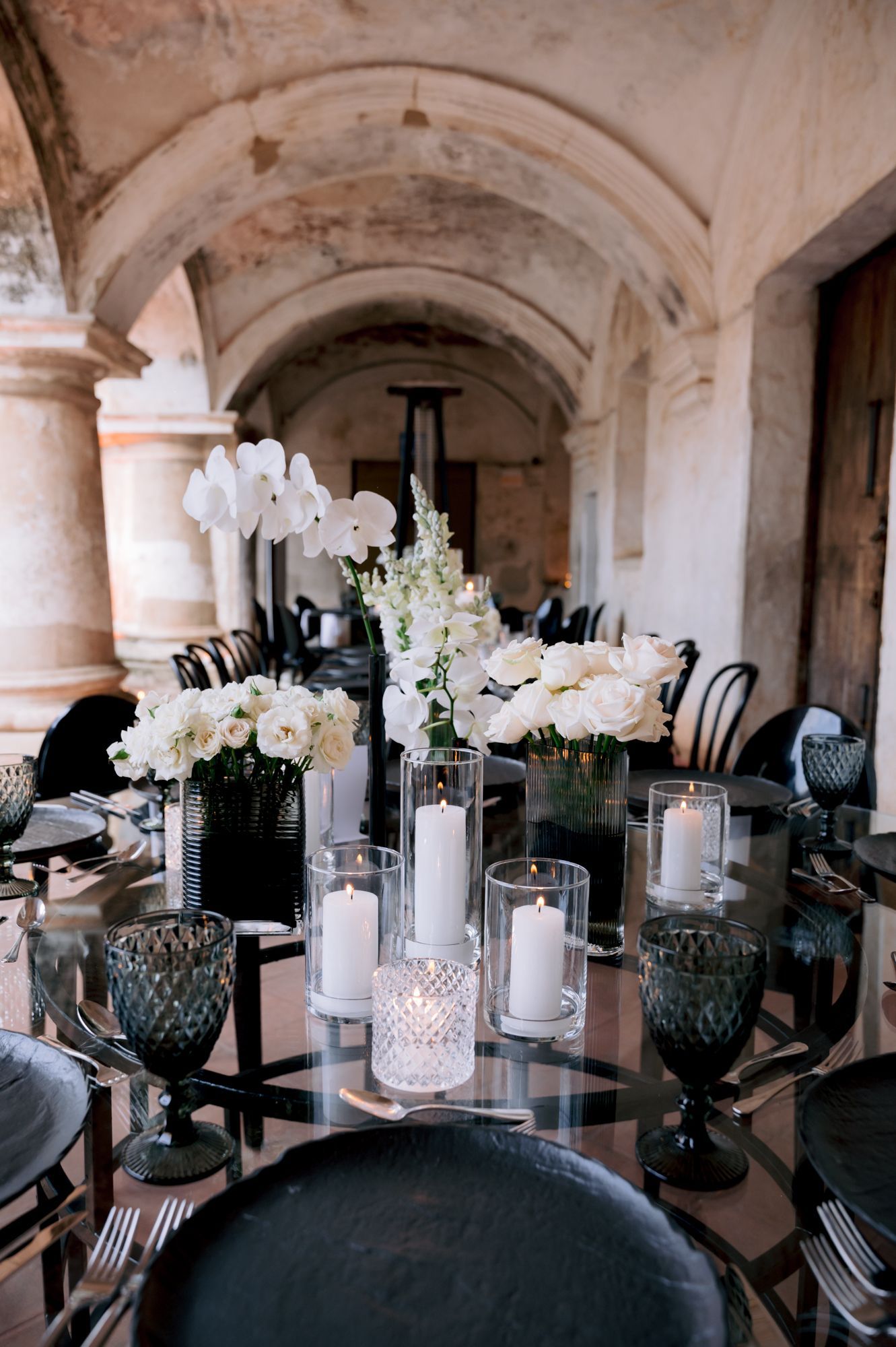 Luxurious wedding table setting with white orchids, candles, and blue glassware under arched ceiling.
