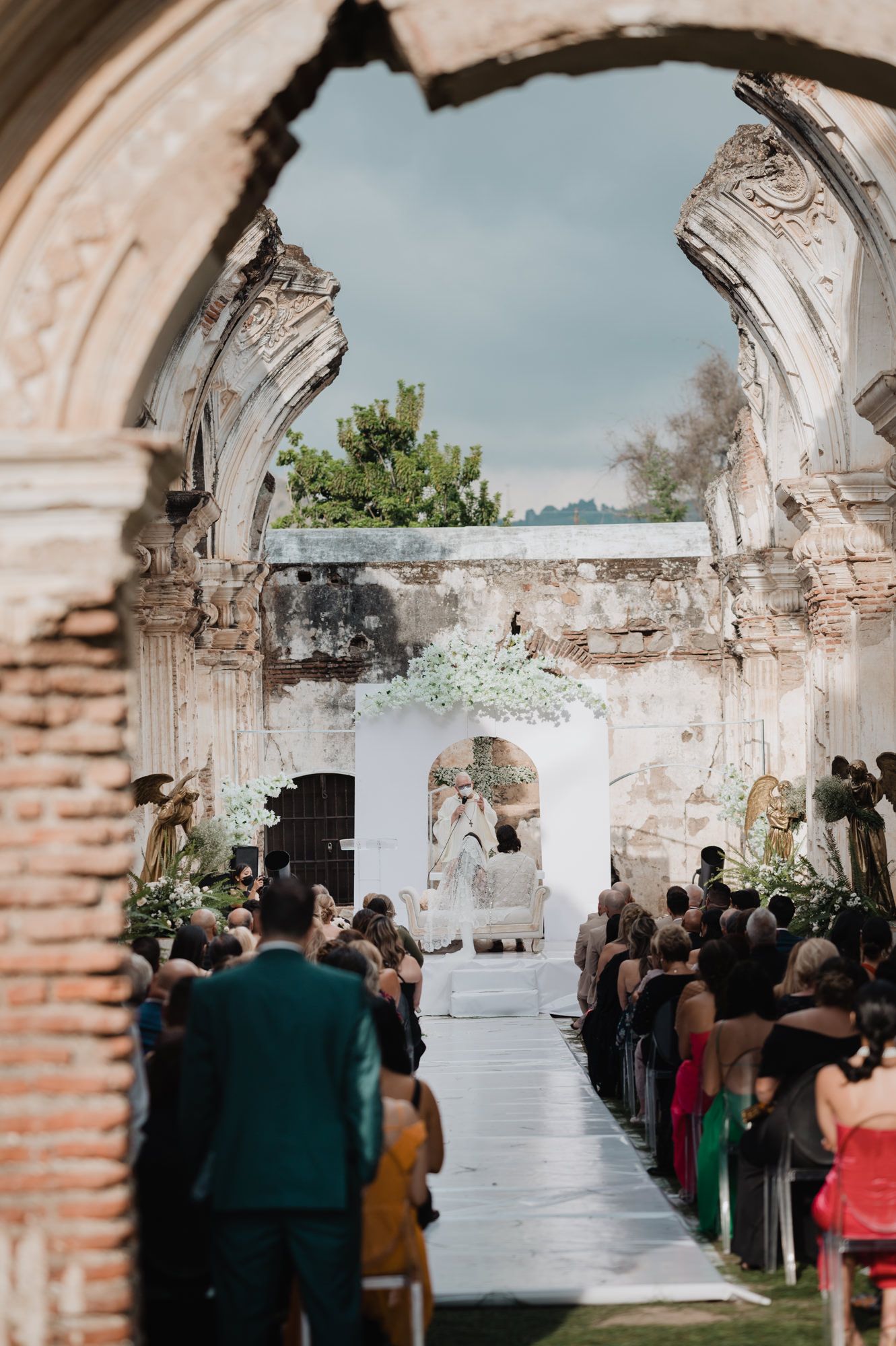 Wedding ceremony setup in an elegant stone archway venue with white floral decorations and seated guests.