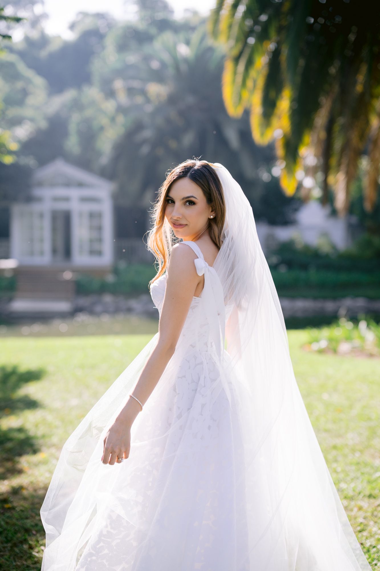 A bride in a flowing white wedding dress and veil poses gracefully in a garden setting with palm trees.