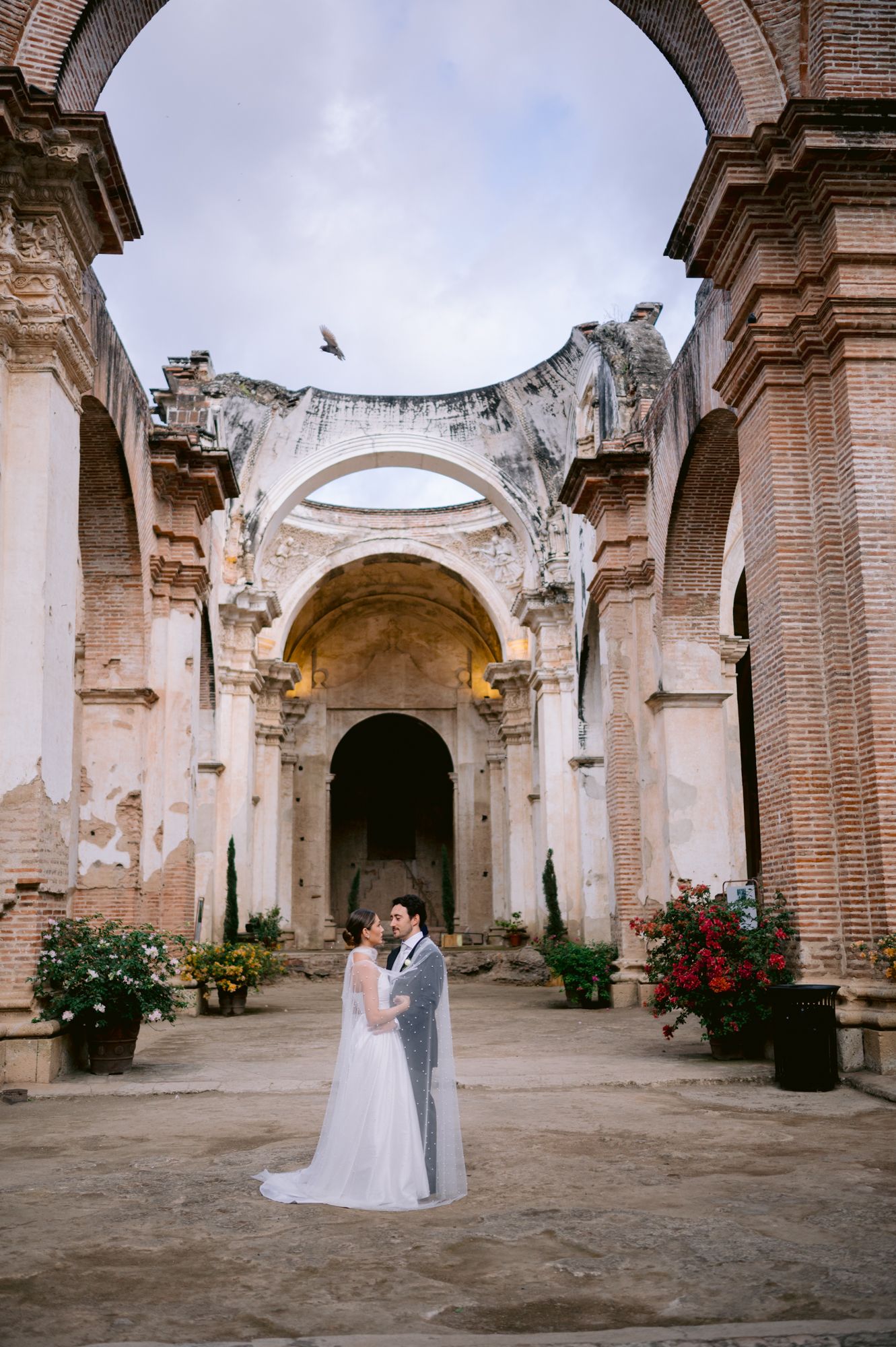 Wedding couple stands in romantic ruins of ancient church with exposed brick archways.