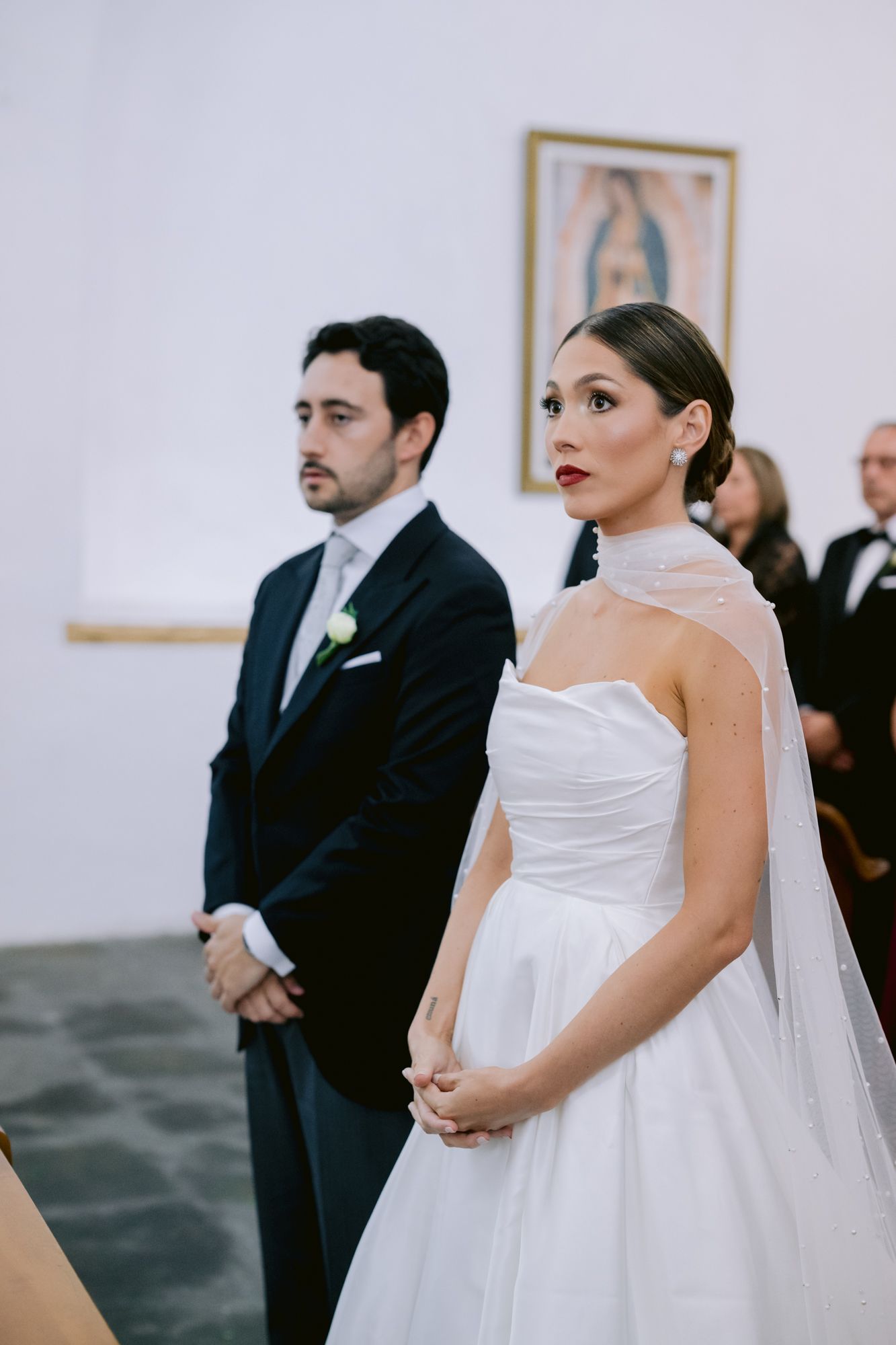 Bride in elegant white gown and groom in black suit stand at altar during traditional church ceremony.