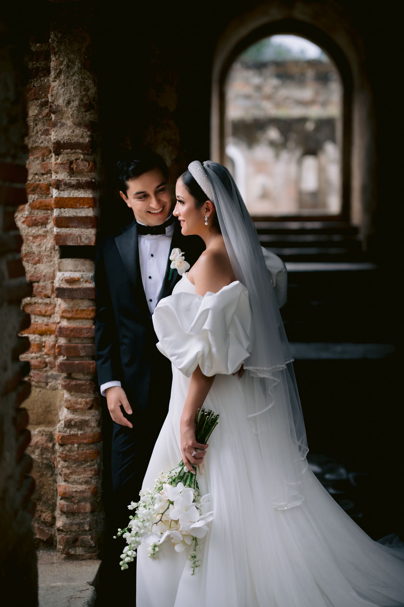 An elegant wedding portrait under a brick archway with a white floral bouquet.