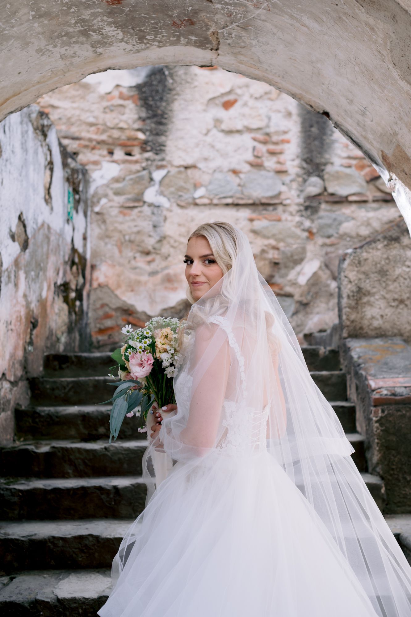 Bride in flowing white gown poses on historic stone steps with elegant bouquet.