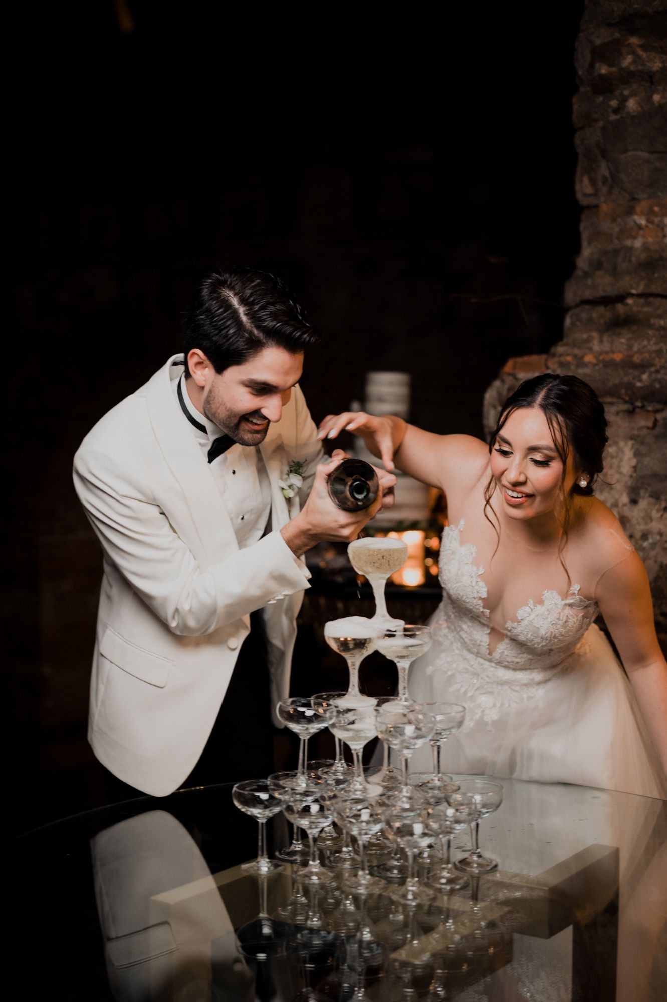 Couple pouring champagne into a tower of glasses during their wedding celebration.
