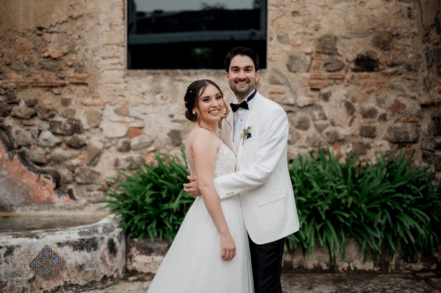 Elegant bride in lace gown and groom in white tuxedo pose against rustic stone wall.
