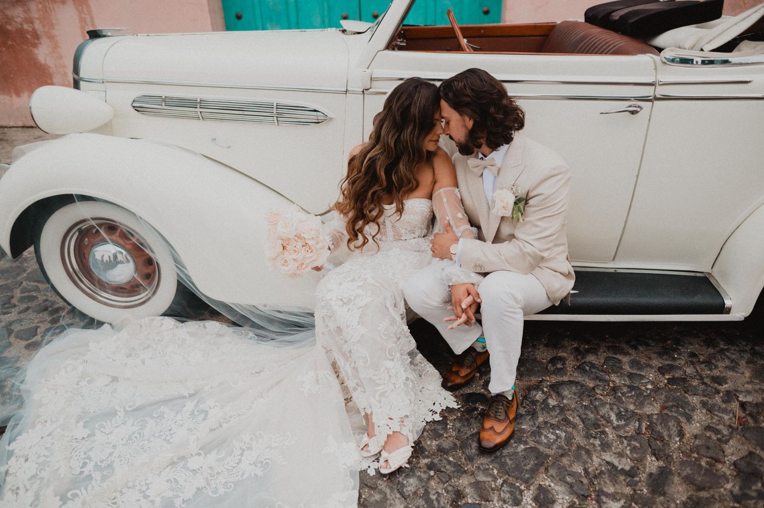 A romantic couple in wedding attire sits together against a classic white vintage car.