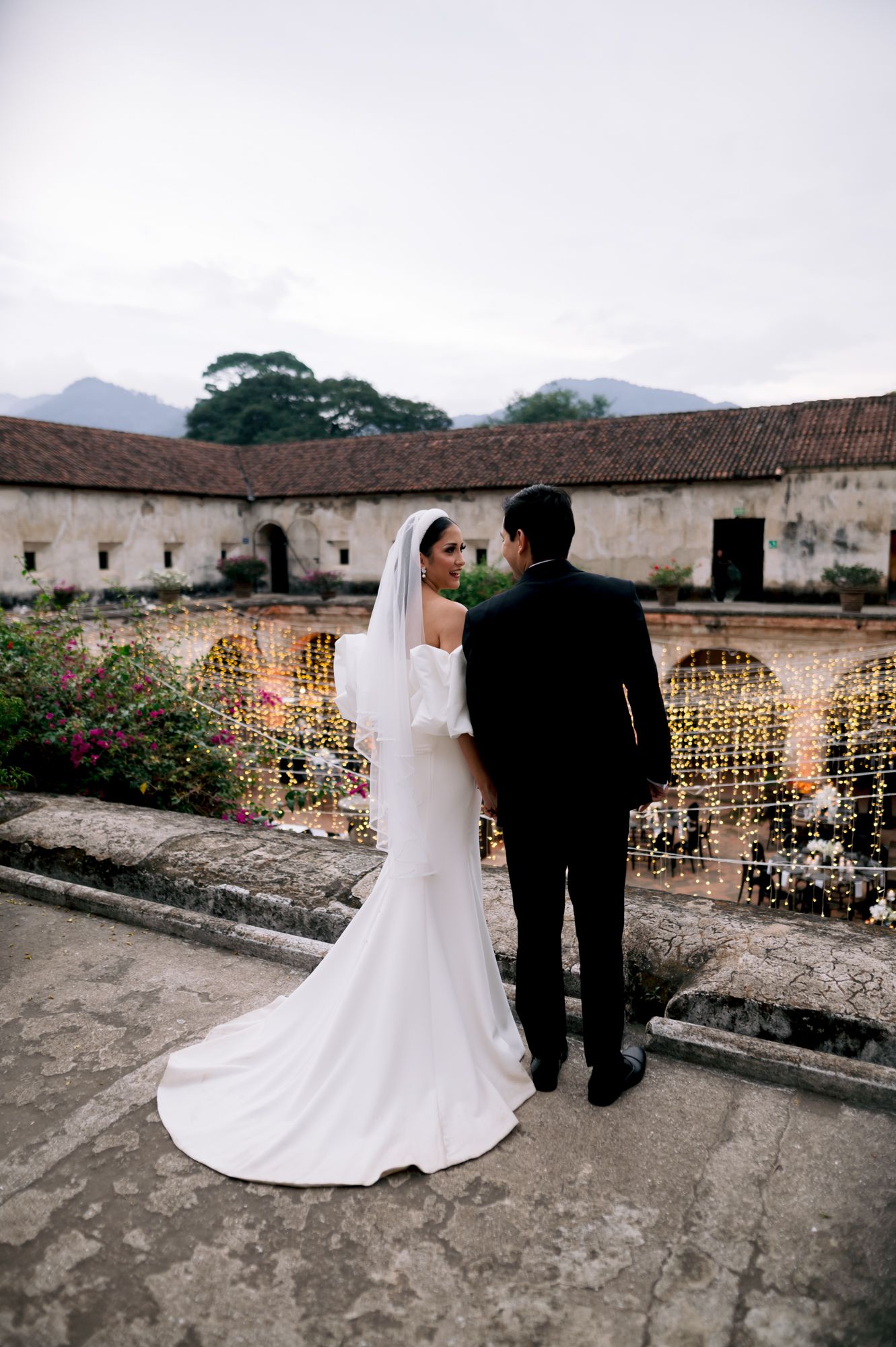 Wedding couple in formal attire overlooking historic courtyard with string lights and mountain backdrop.