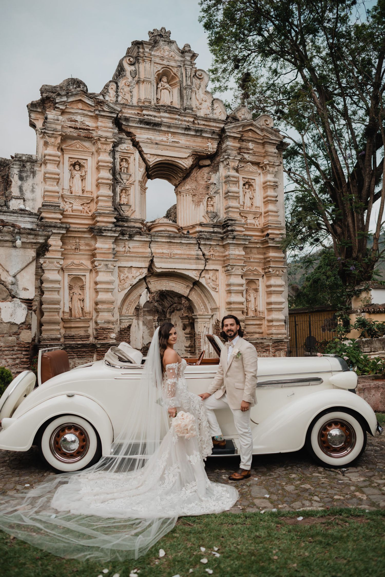 Wedding couple poses with vintage white car in front of ornate colonial ruins.