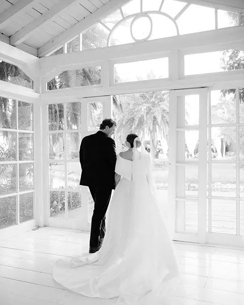 A silhouetted couple in bridal attire stands together in a bright sunlit conservatory with large windows and white decor.