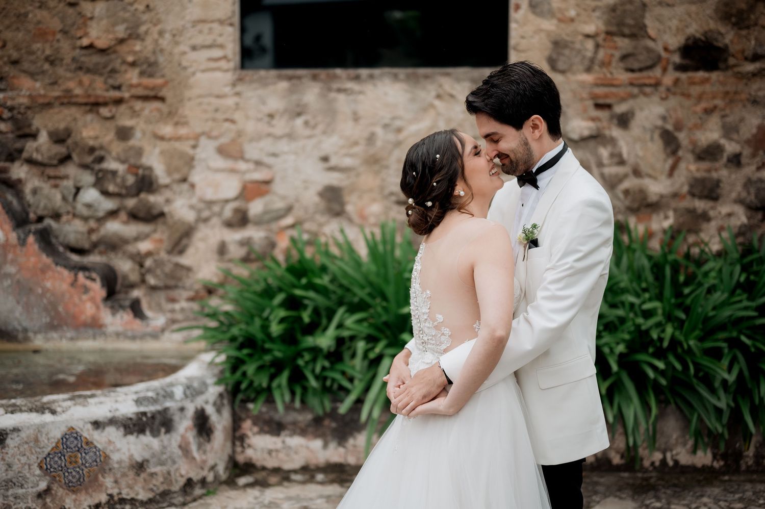 Tender wedding moment captured against historic stone wall and tropical foliage.