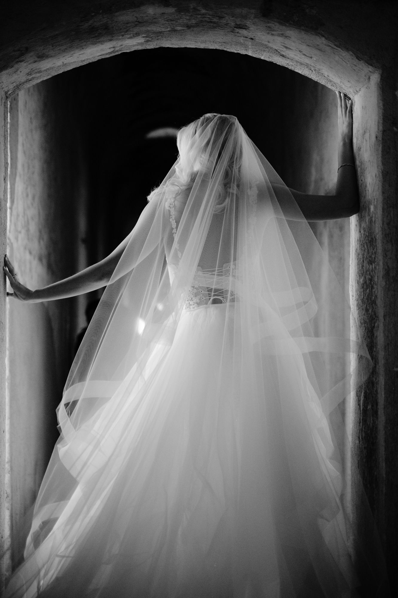 Artistic black and white silhouette of a bride with flowing veil in a stone passage.