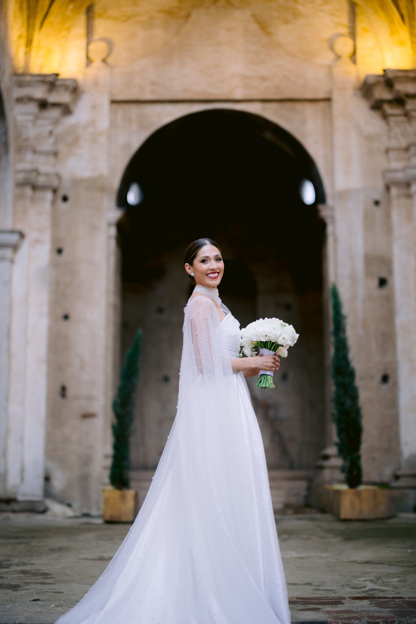Bride in elegant white gown holding bouquet poses in front of ornate church architecture.