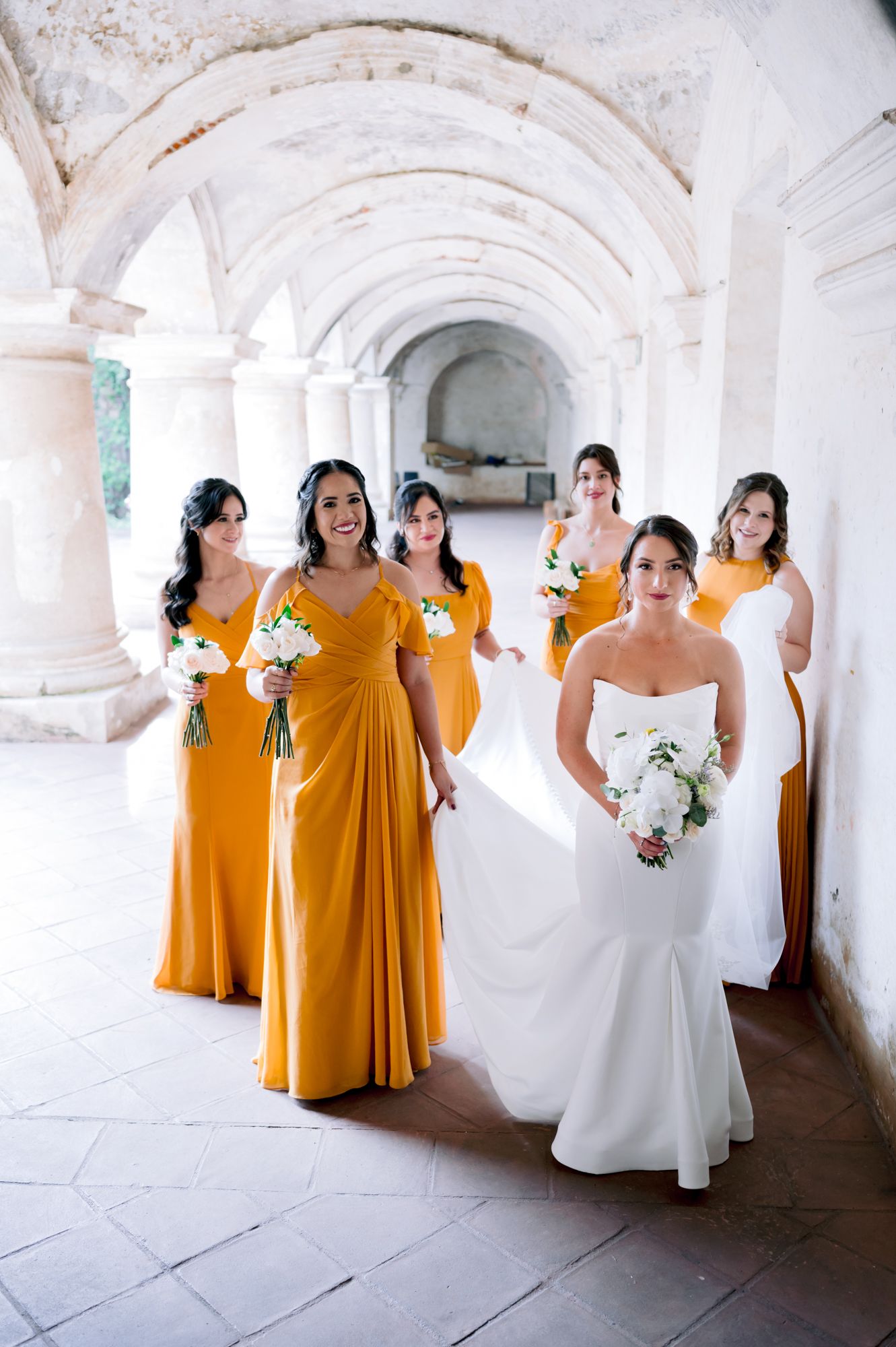 Wedding party photo featuring bridesmaids in mustard gowns under white arched colonnade.