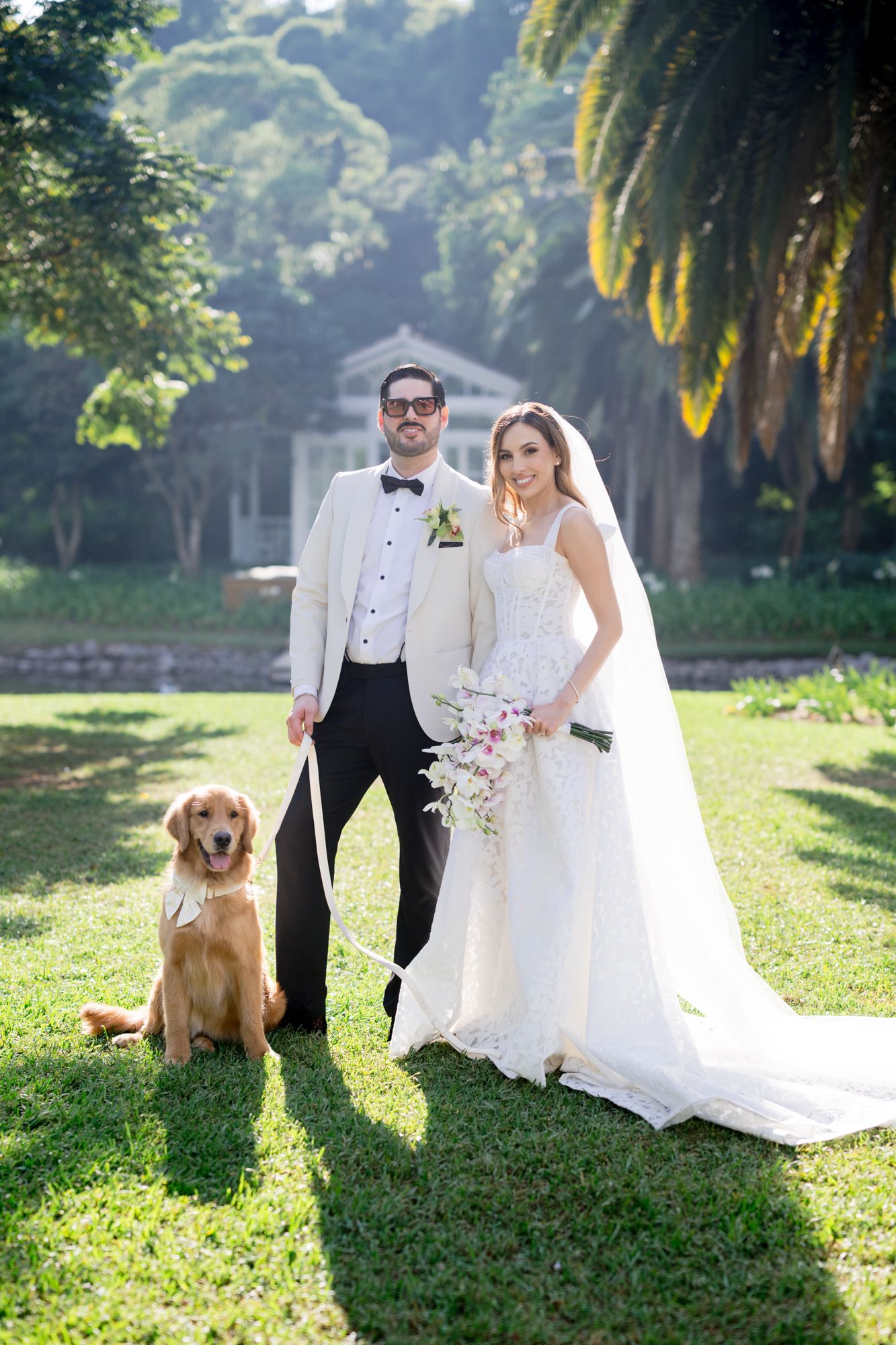 A wedding couple poses with their golden retriever on a sunny lawn with palm trees and a white building behind them.