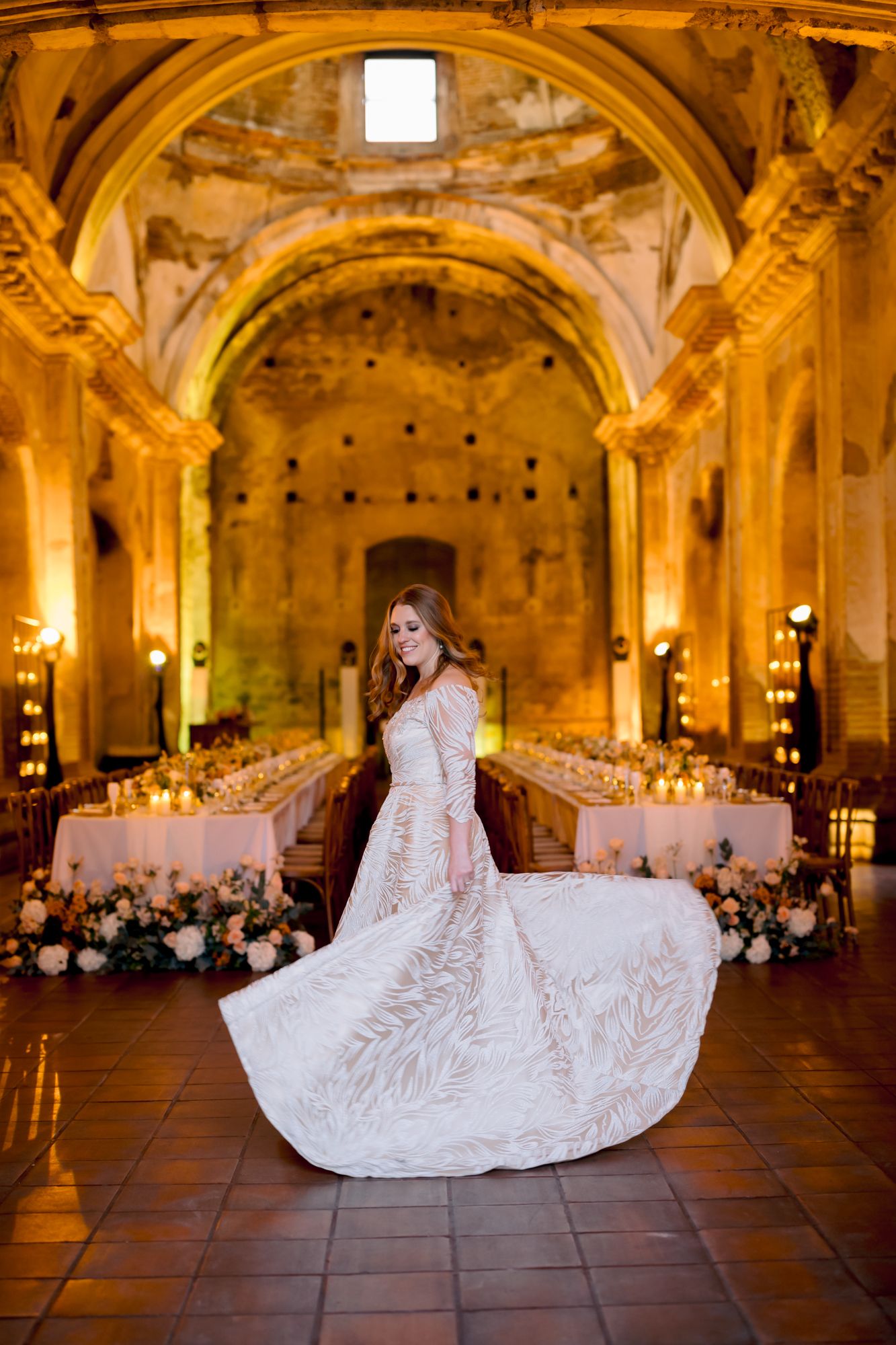 Elegant wedding gown swirling in a candlelit historic chapel with long banquet tables and floral arrangements.