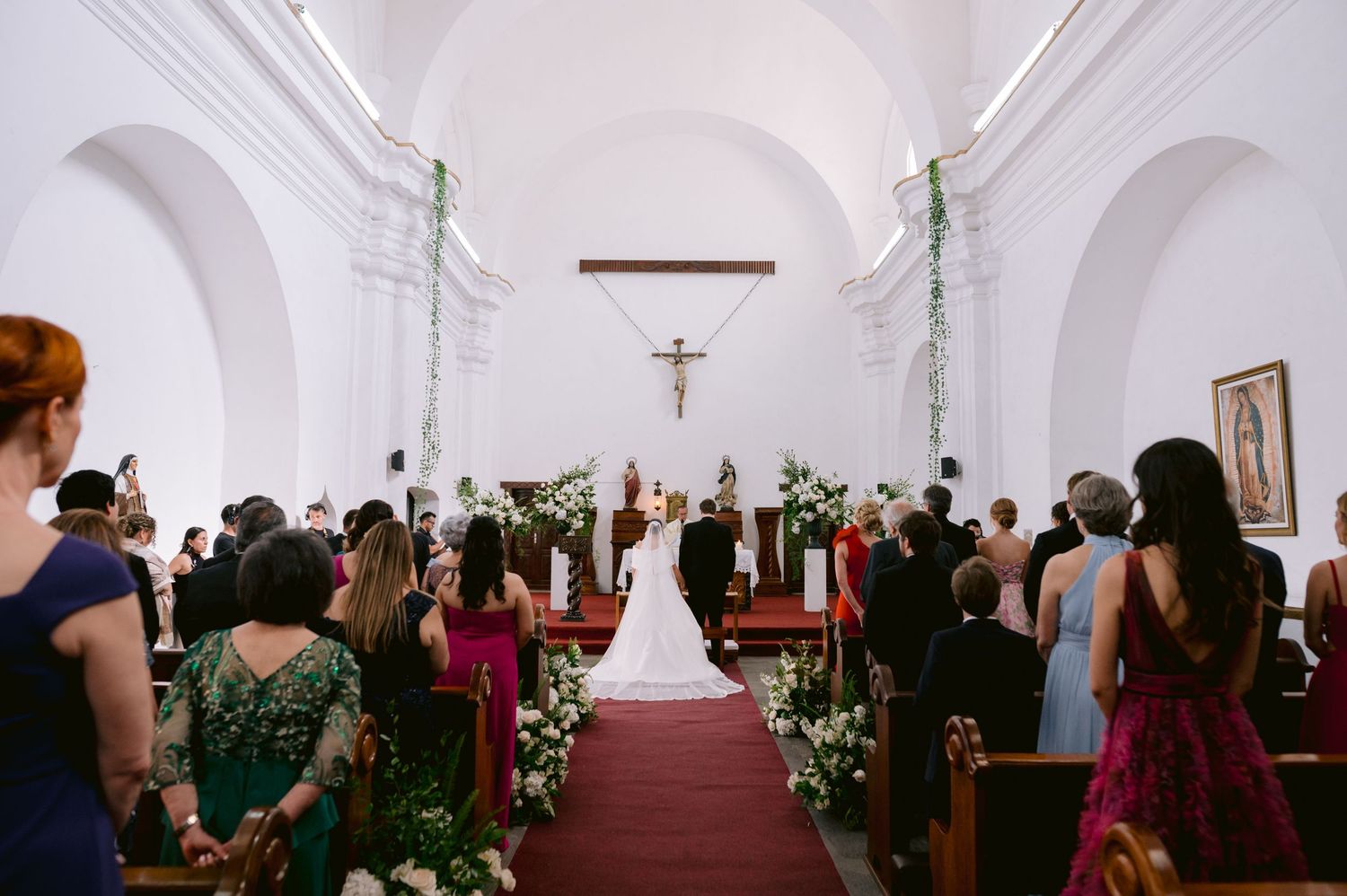 Wedding ceremony in progress inside a white church with guests standing along the red-carpeted aisle.