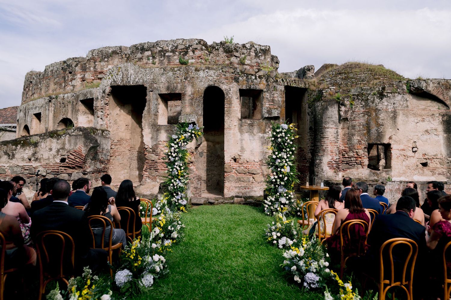 Wedding ceremony venue with floral arch decorations at ancient stone ruins with guests seated.