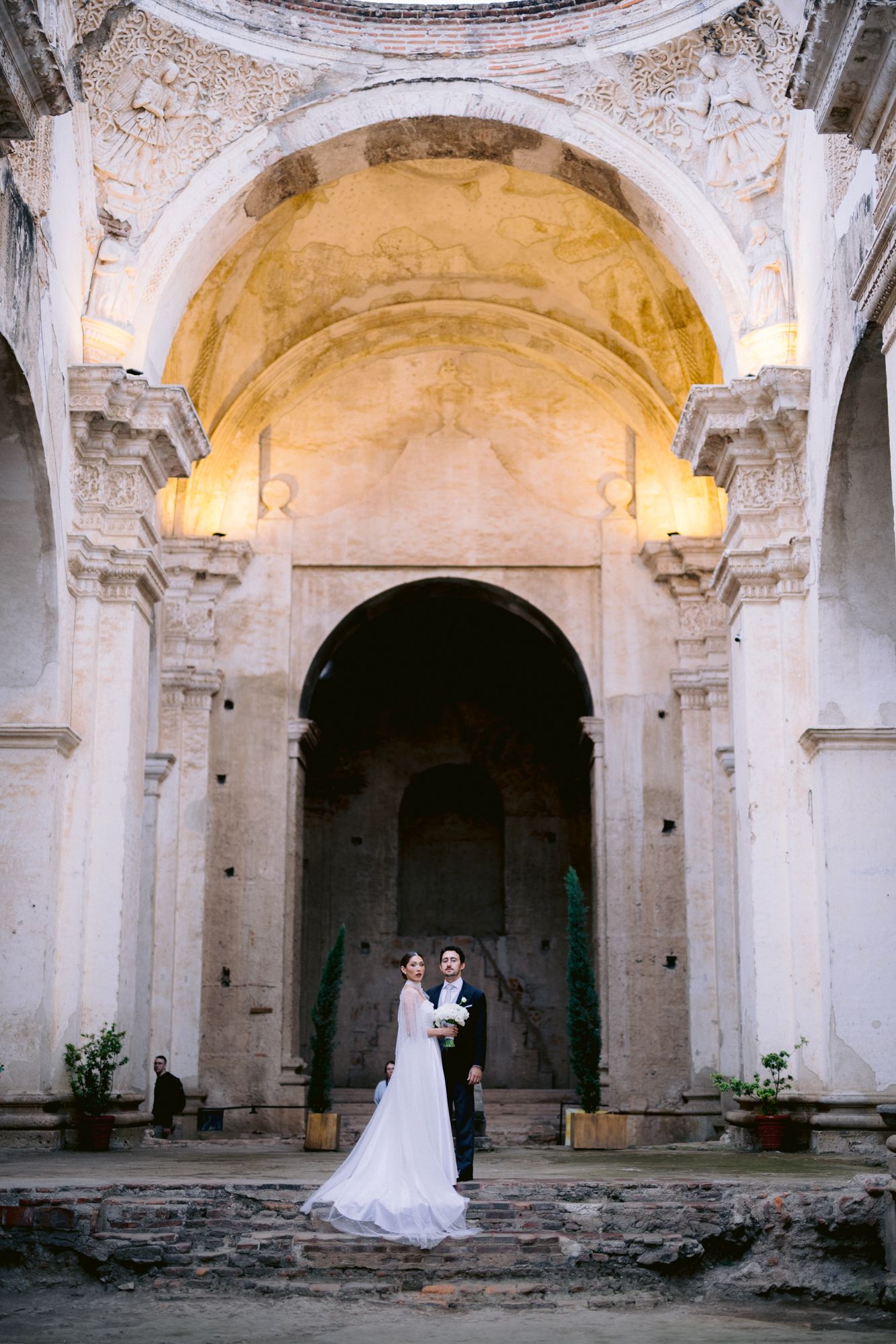 Wedding couple stands beneath majestic church dome and arches in romantic ceremony setting.