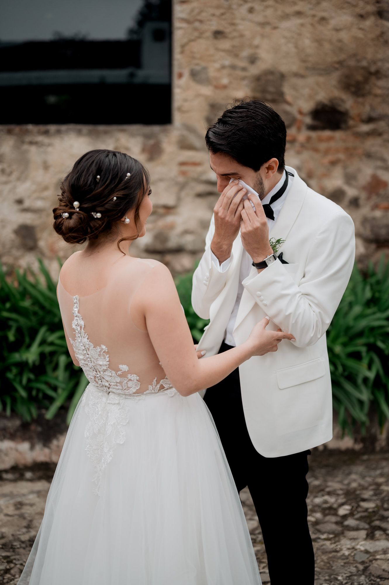 Tearful wedding moment between couple in elegant attire by rustic wall.