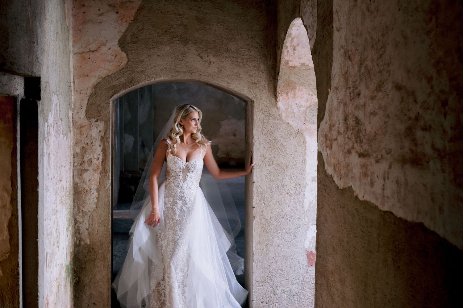 Bride wearing an elegant lace gown stands framed by weathered stone architecture.
