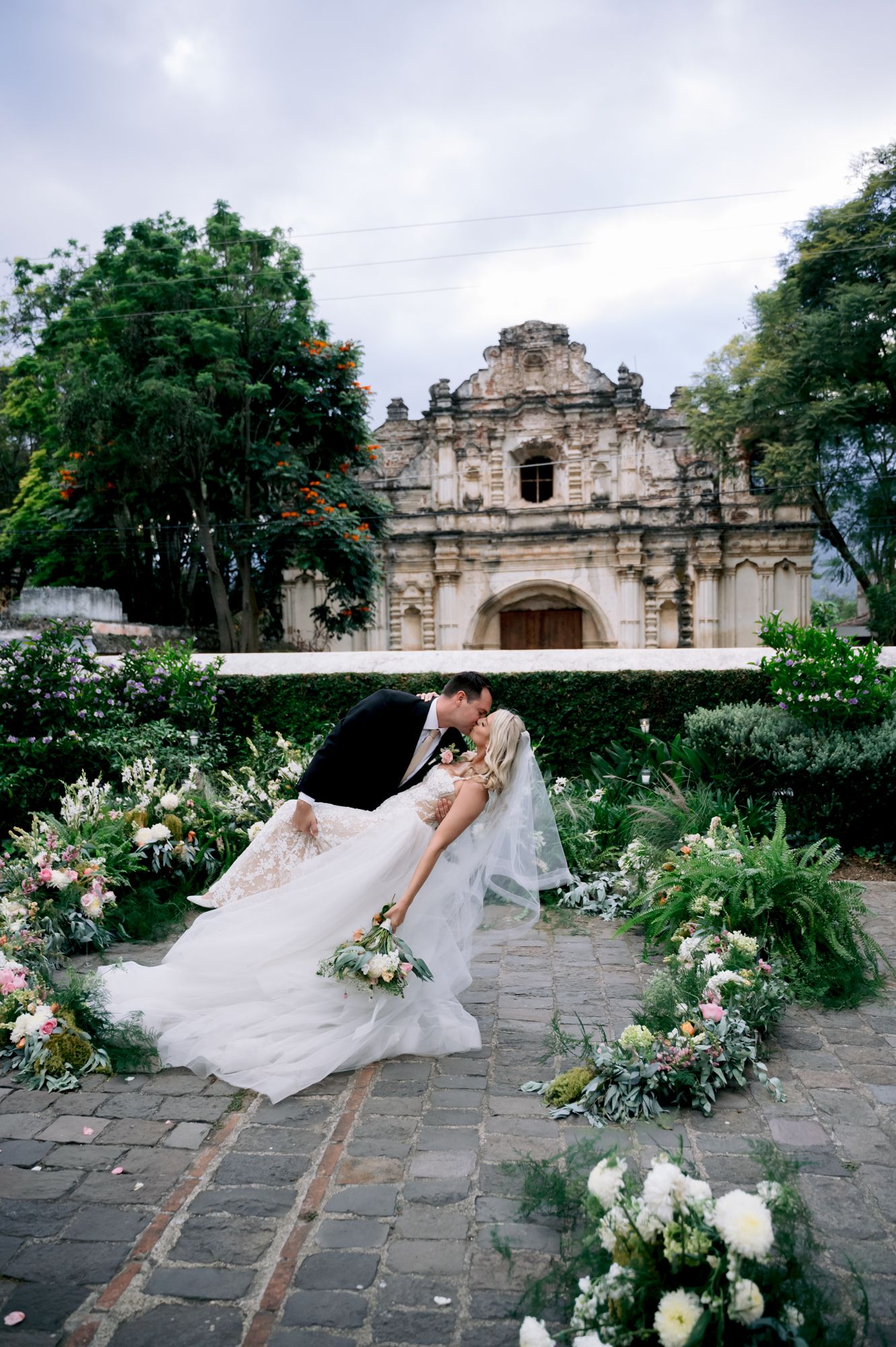 Wedding couple embraces in garden with colonial architecture and floral arrangements.