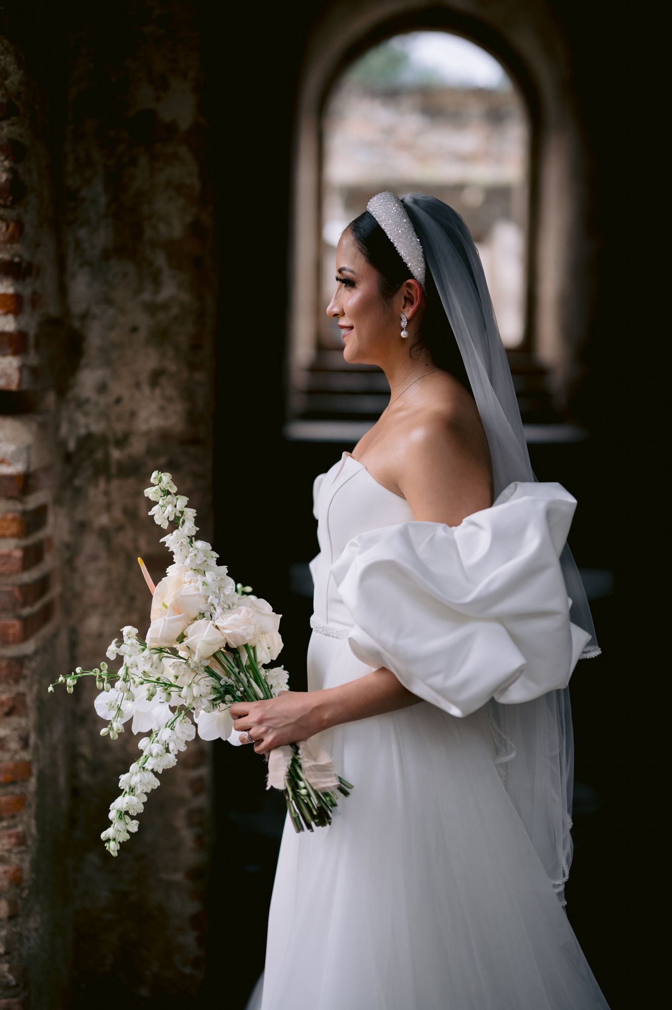 An elegant bride in an off-shoulder gown holds a white floral bouquet against rustic brick walls.