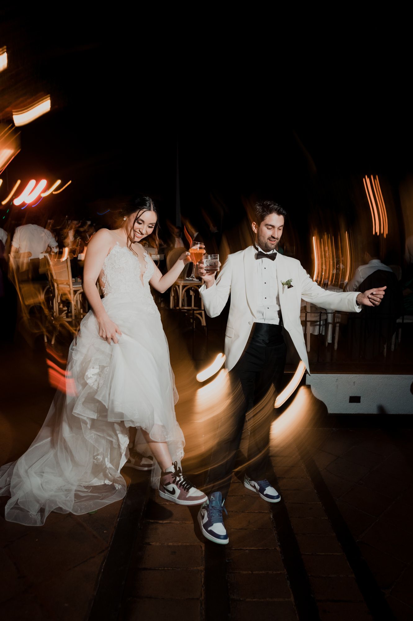 Wedding couple dancing energetically in formal attire and sneakers under ambient lighting.