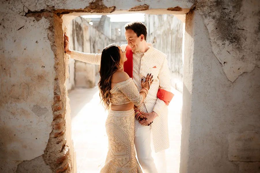 Two people in light colored clothing embrace in a romantic pose framed by a rustic stone doorway filled with sunlight.