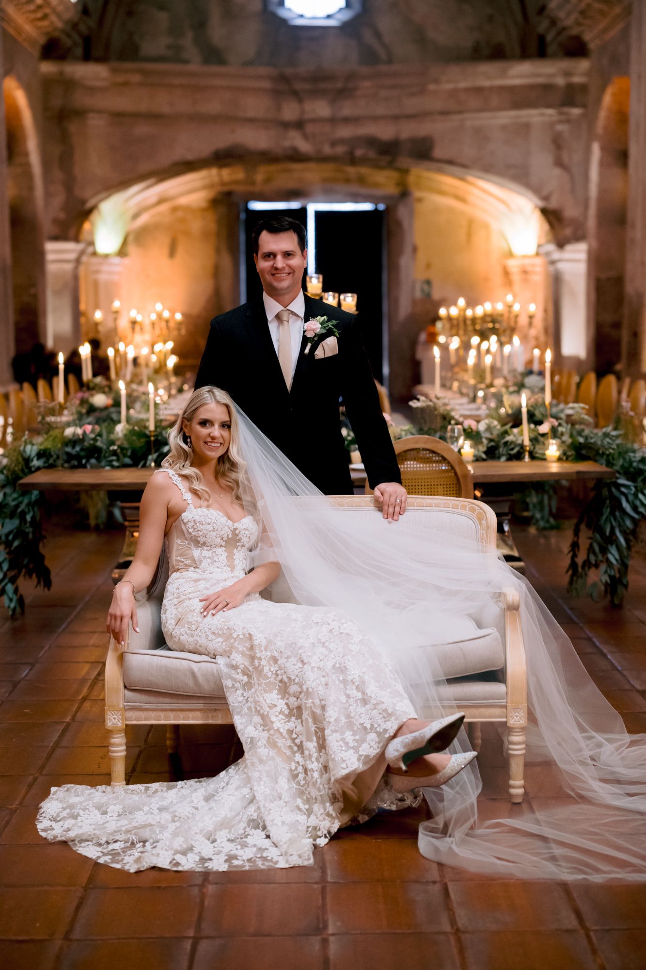 A couple poses in an ornate stone venue with candles and greenery decorating the elegant reception space.
