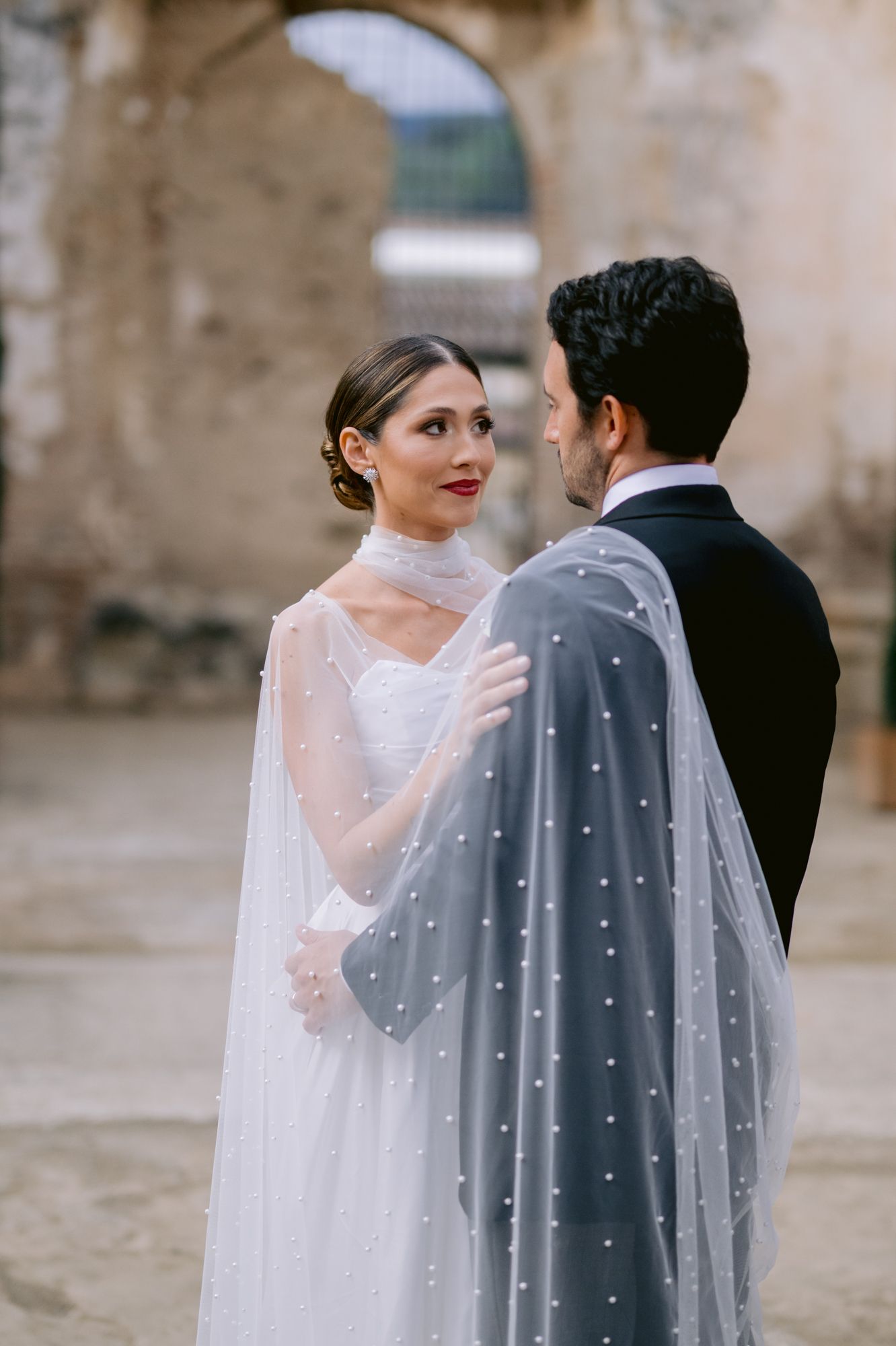 Close-up of wedding couple wrapped in pearl-studded veil against historic backdrop.