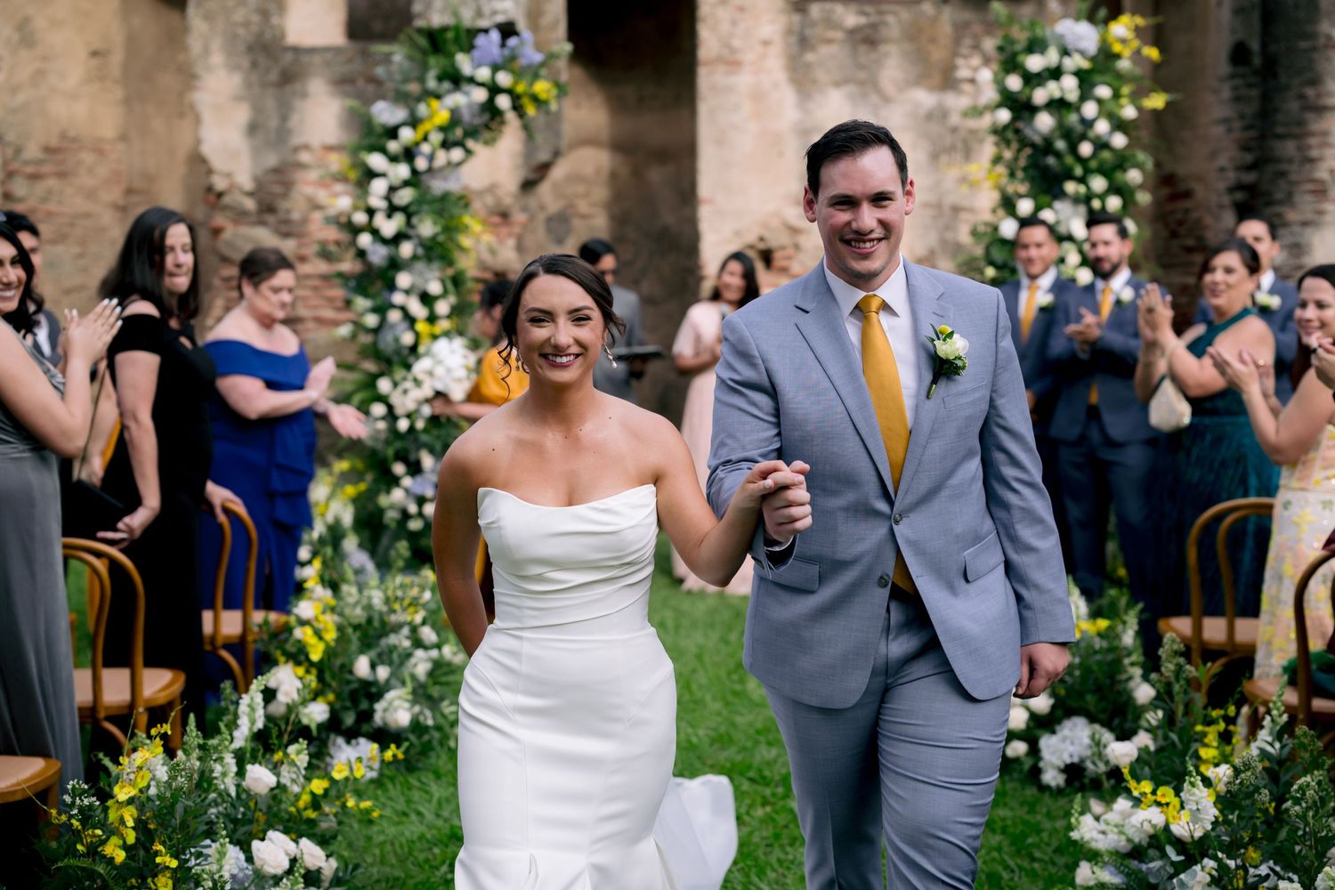 Newlyweds walk down an outdoor aisle with floral archways and decorations at a garden wedding ceremony.