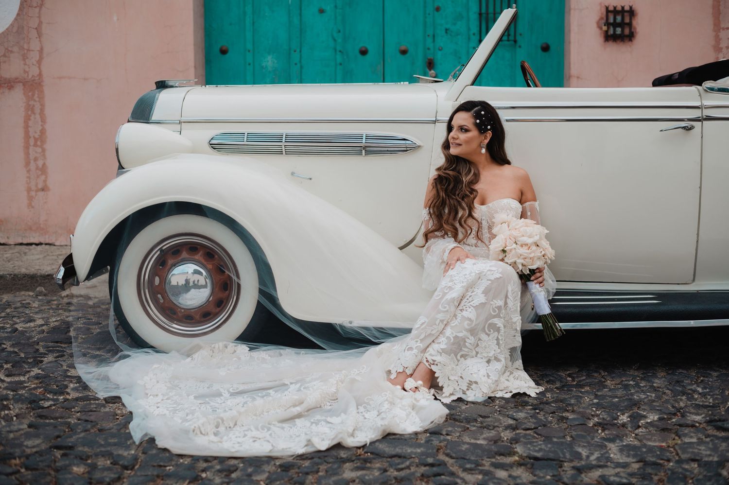 Wedding couple poses beside vintage white car against turquoise door and pink wall backdrop.