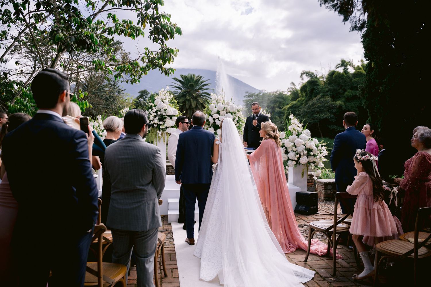 Wedding ceremony guests gather outdoors under trees with mountains visible in the background.