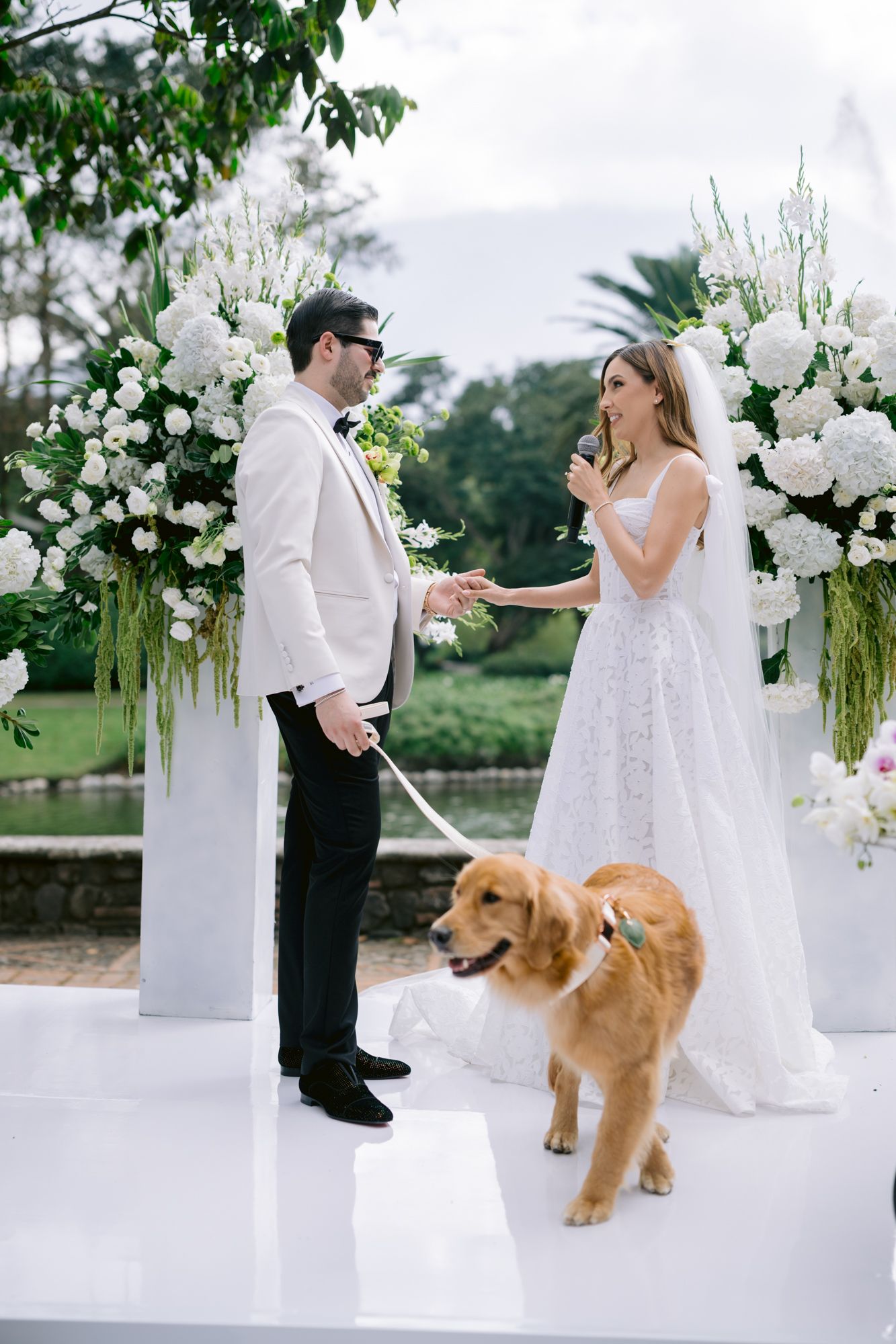 A couple shares a toast at an elegant outdoor wedding ceremony with white floral arrangements and a golden retriever.