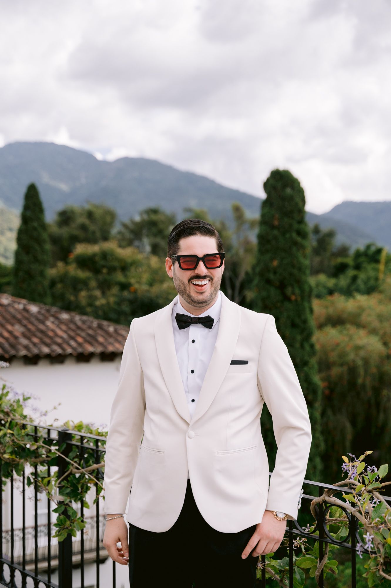 A person wearing a white tuxedo jacket poses against a scenic mountain backdrop with trees and cloudy skies.