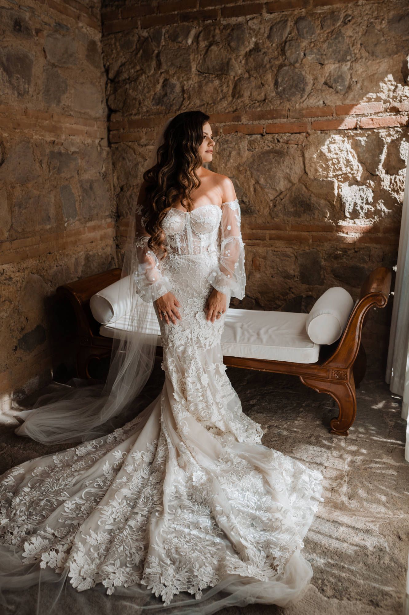 A stunning lace wedding dress with off-shoulder sleeves flows across a rustic stone floor in an antique setting.