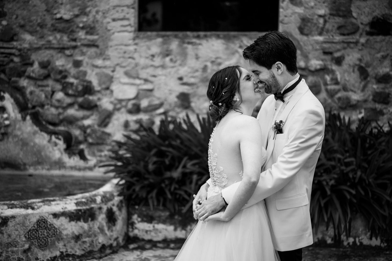 A romantic black and white wedding photo of a couple sharing a tender moment against a stone wall.