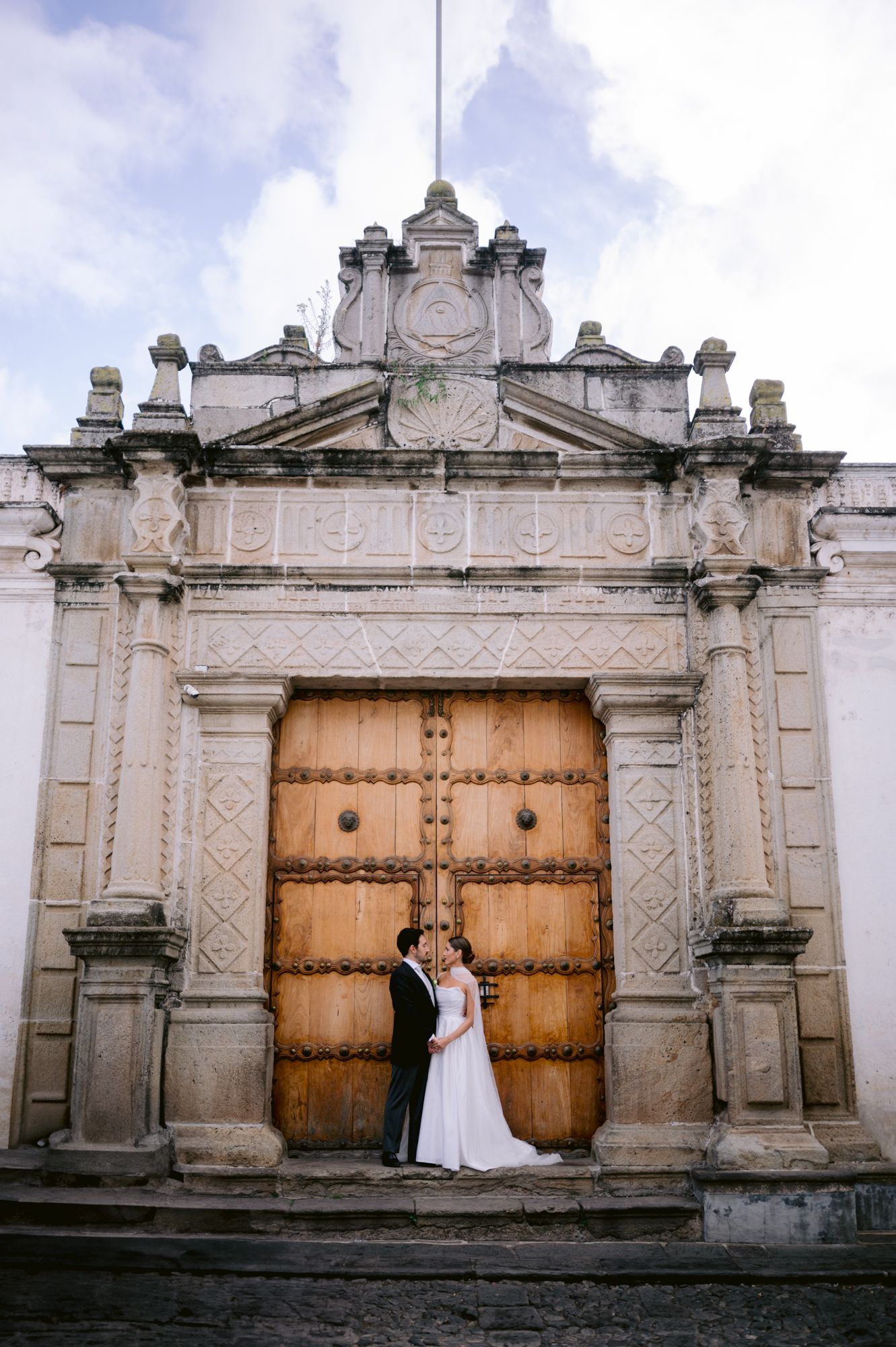 Newlyweds pose before massive ornate wooden doors of historic stone church entrance.