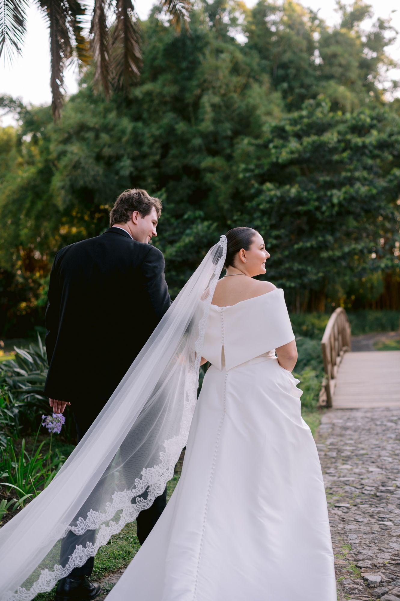 A wedding couple walking along a garden path with a dramatic lace-trimmed veil trailing behind.