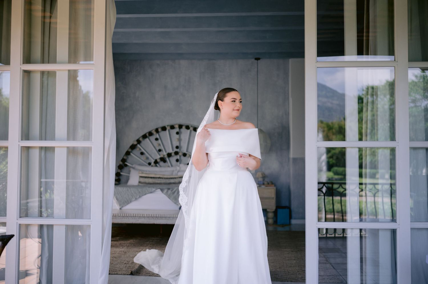 Bride in elegant white dress standing in doorway of bedroom with french doors.