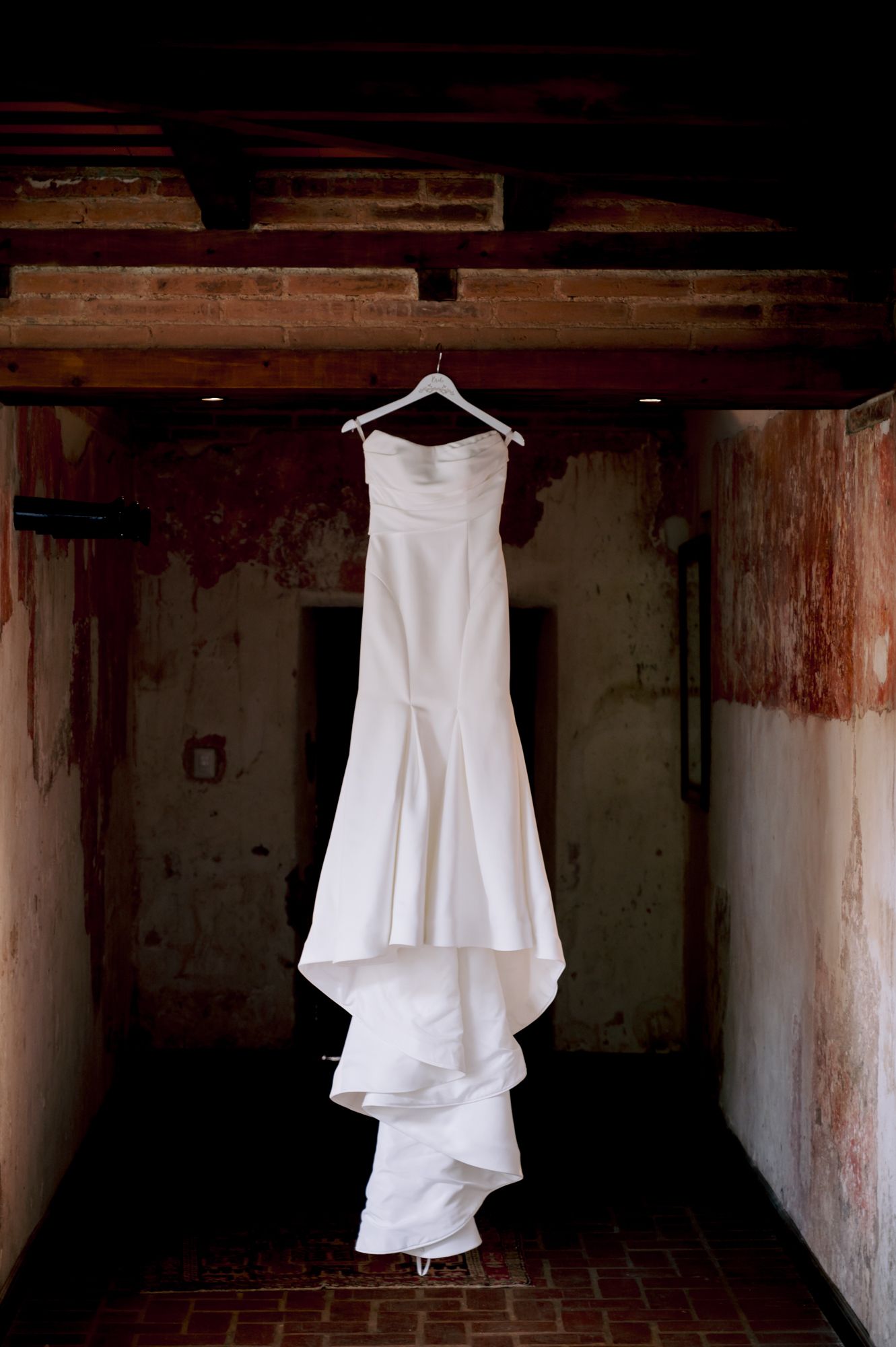 Dramatic photo of white wedding dress hanging in dark rustic hallway.