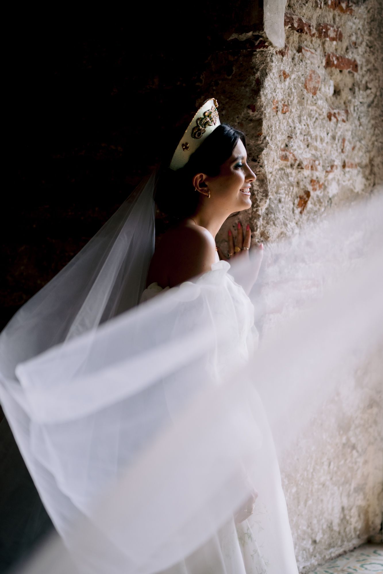 Bride in an ornate headpiece standing near a textured brick wall with flowing veil.
