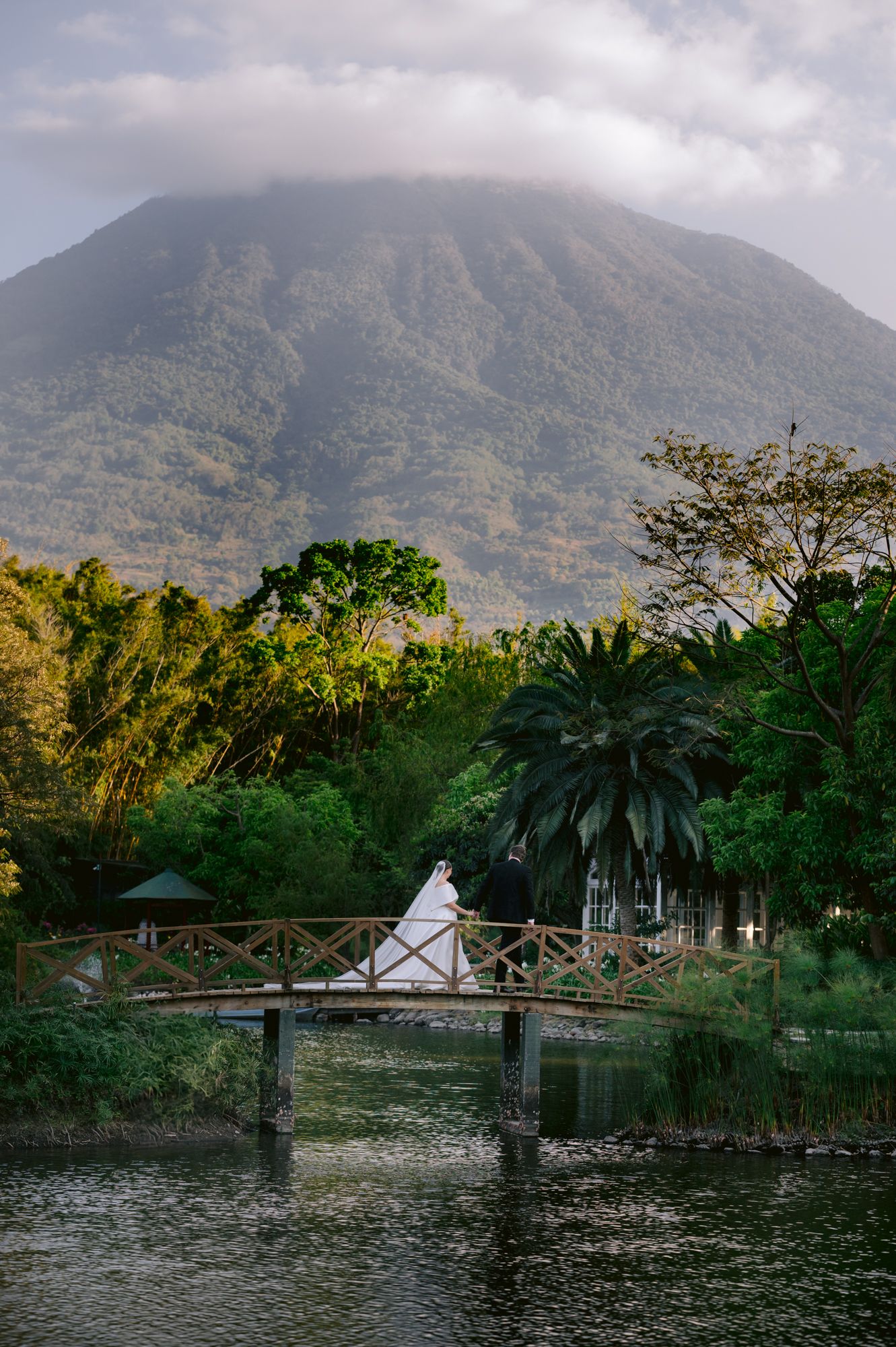 Wedding photo on wooden bridge with volcanic mountain and tropical landscape in background.