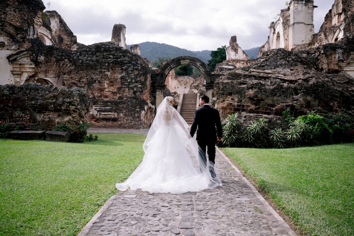 Couple walks along stone path surrounded by ancient ruins and green lawn.