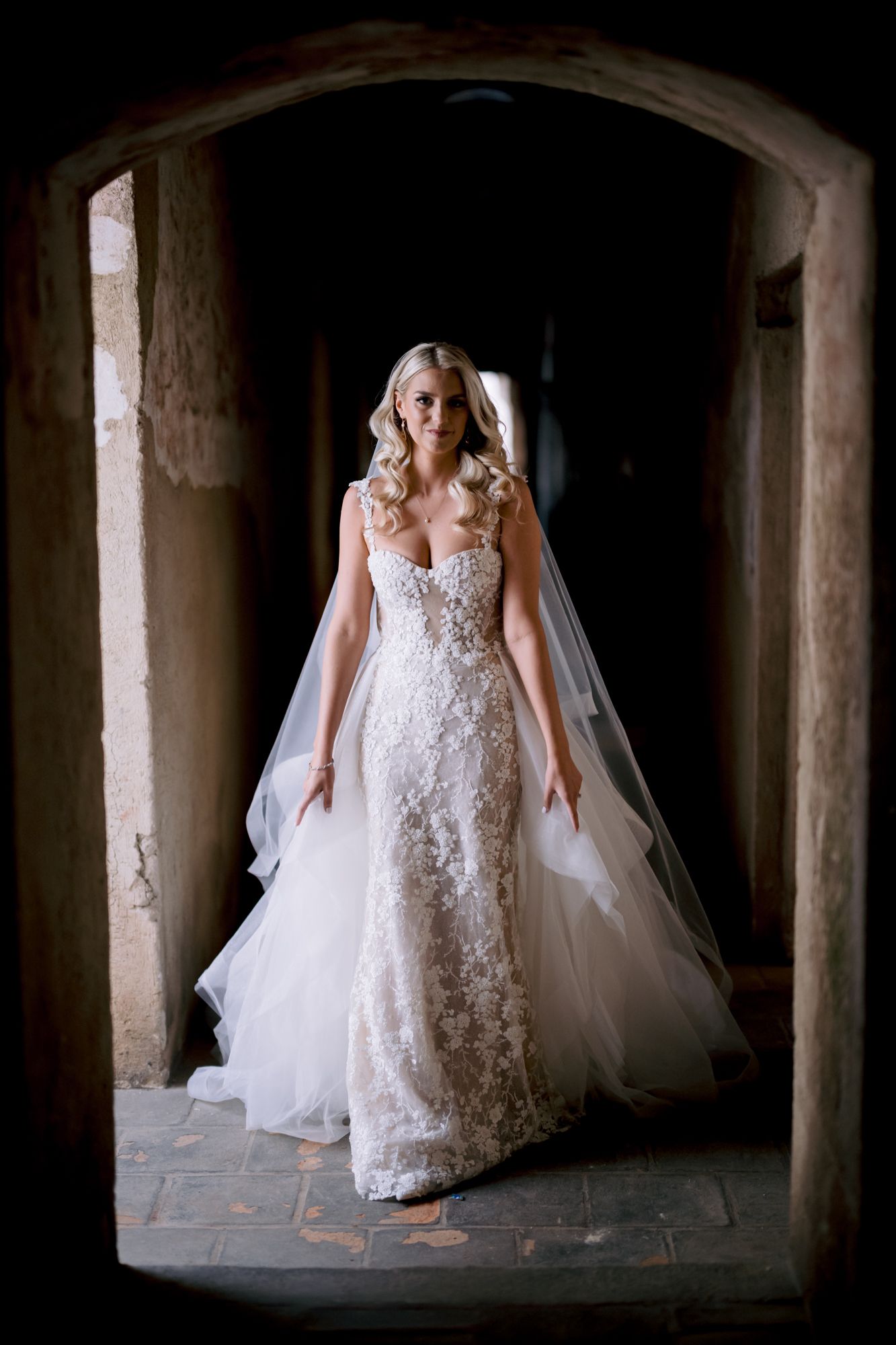 Bride in form-fitting lace wedding dress stands framed by ancient stone archway.