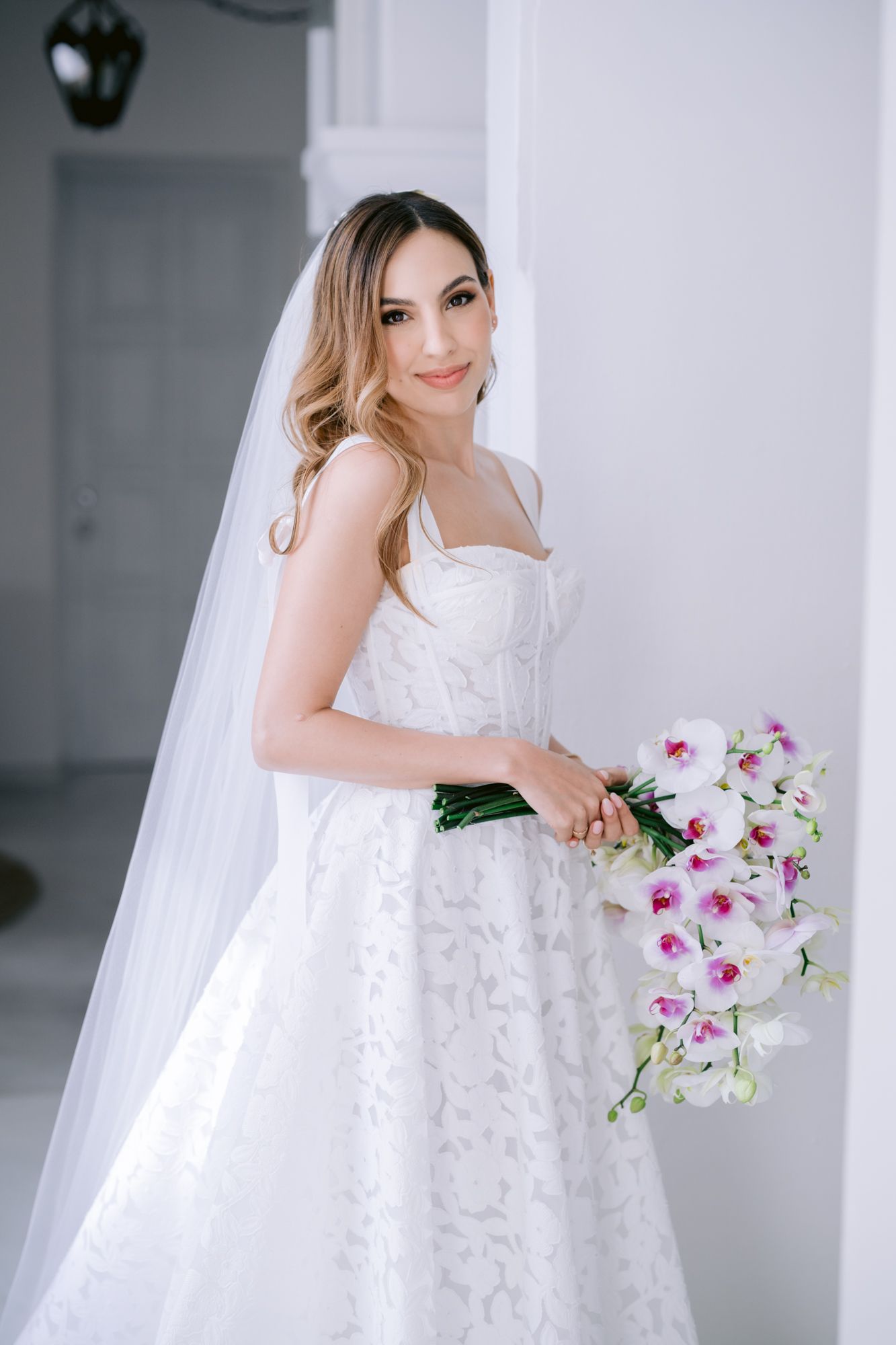 Close-up details of white orchid bouquet and lace wedding dress in bright interior setting.