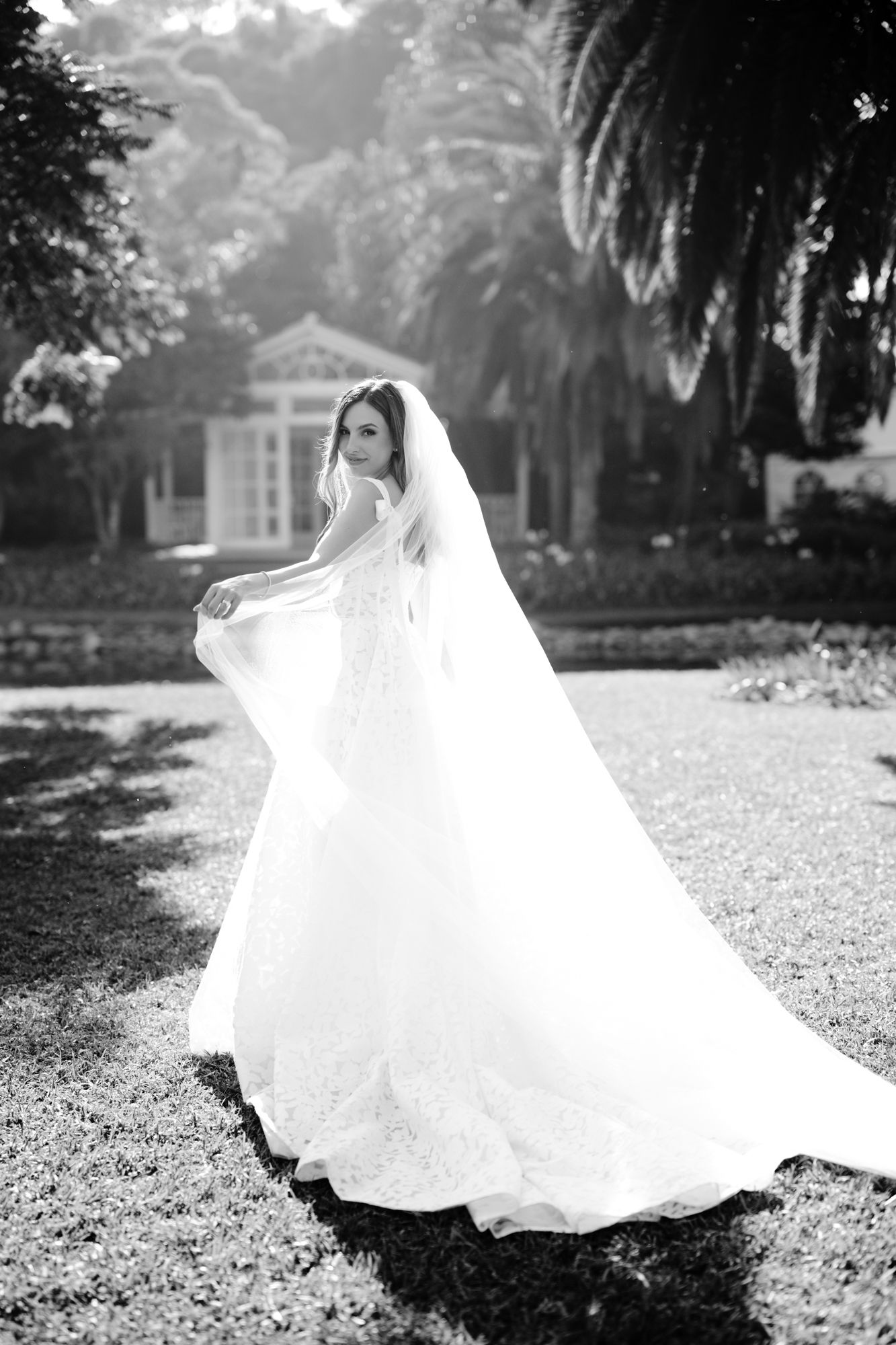 Black and white bridal portrait showing flowing wedding dress and veil in garden with palm trees.