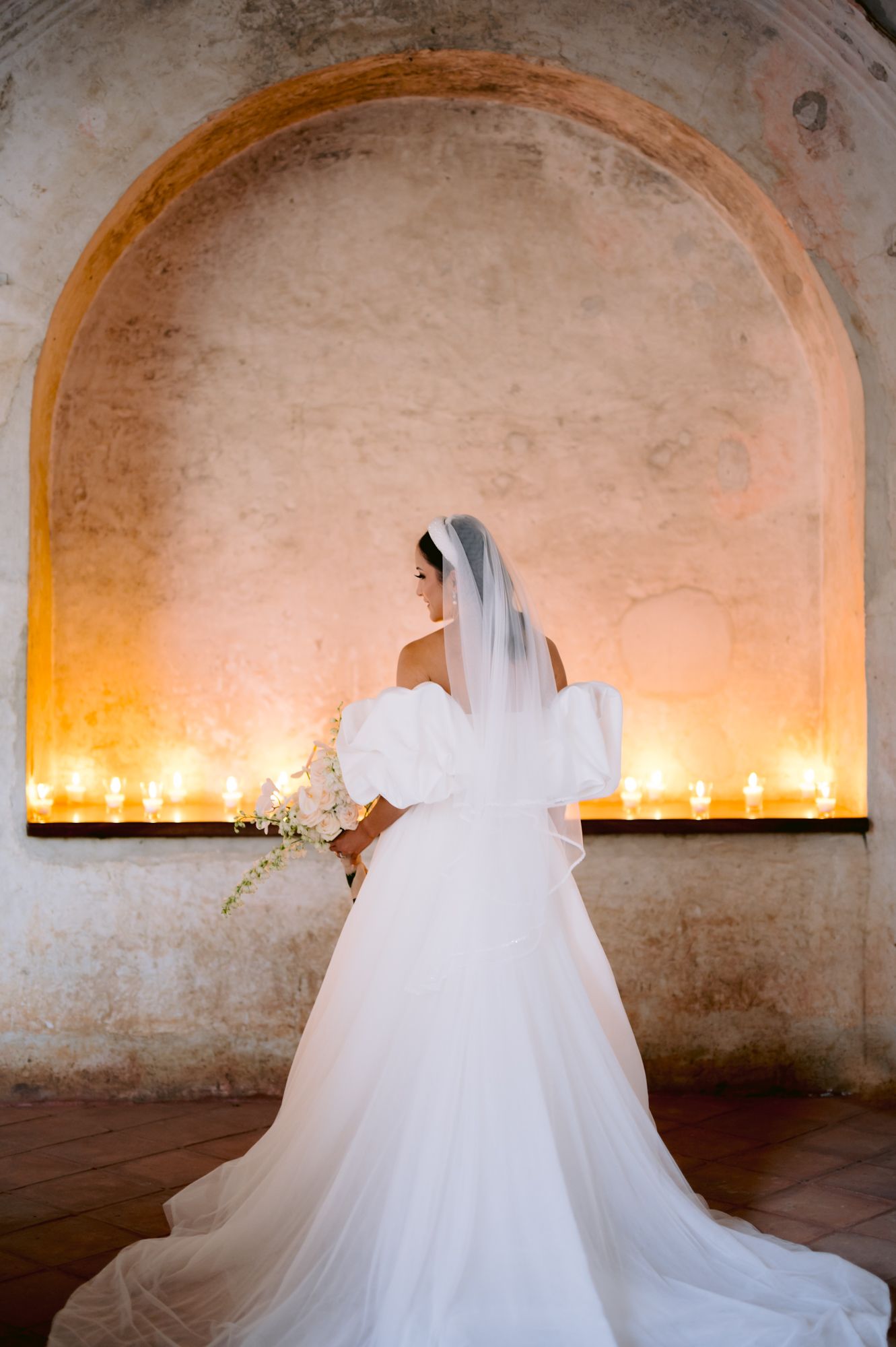 A bride in a flowing white gown stands before an illuminated arch filled with glowing candles.