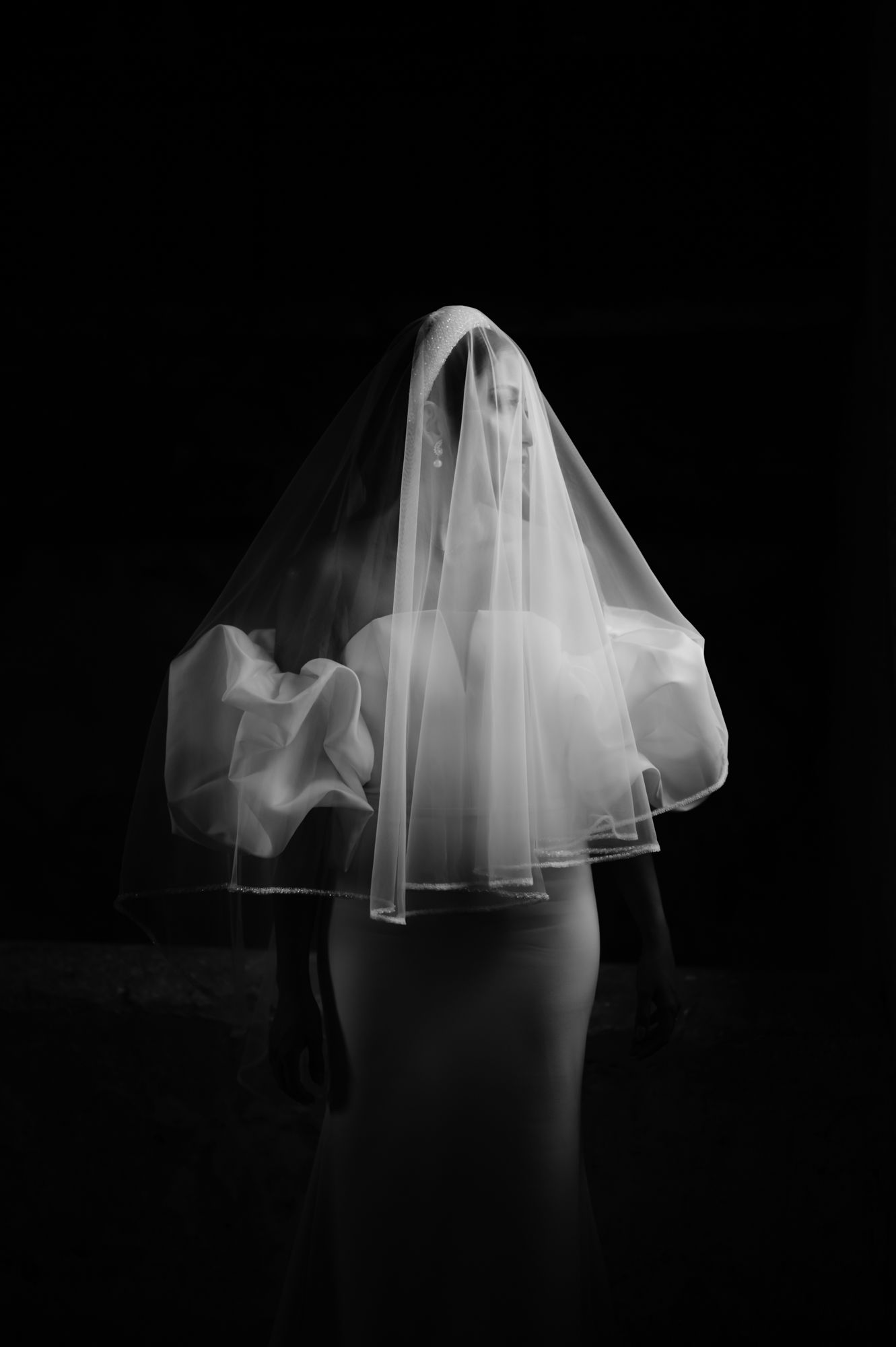 Artistic black and white photo of bride's silhouette with dramatically flowing veil against dark background.